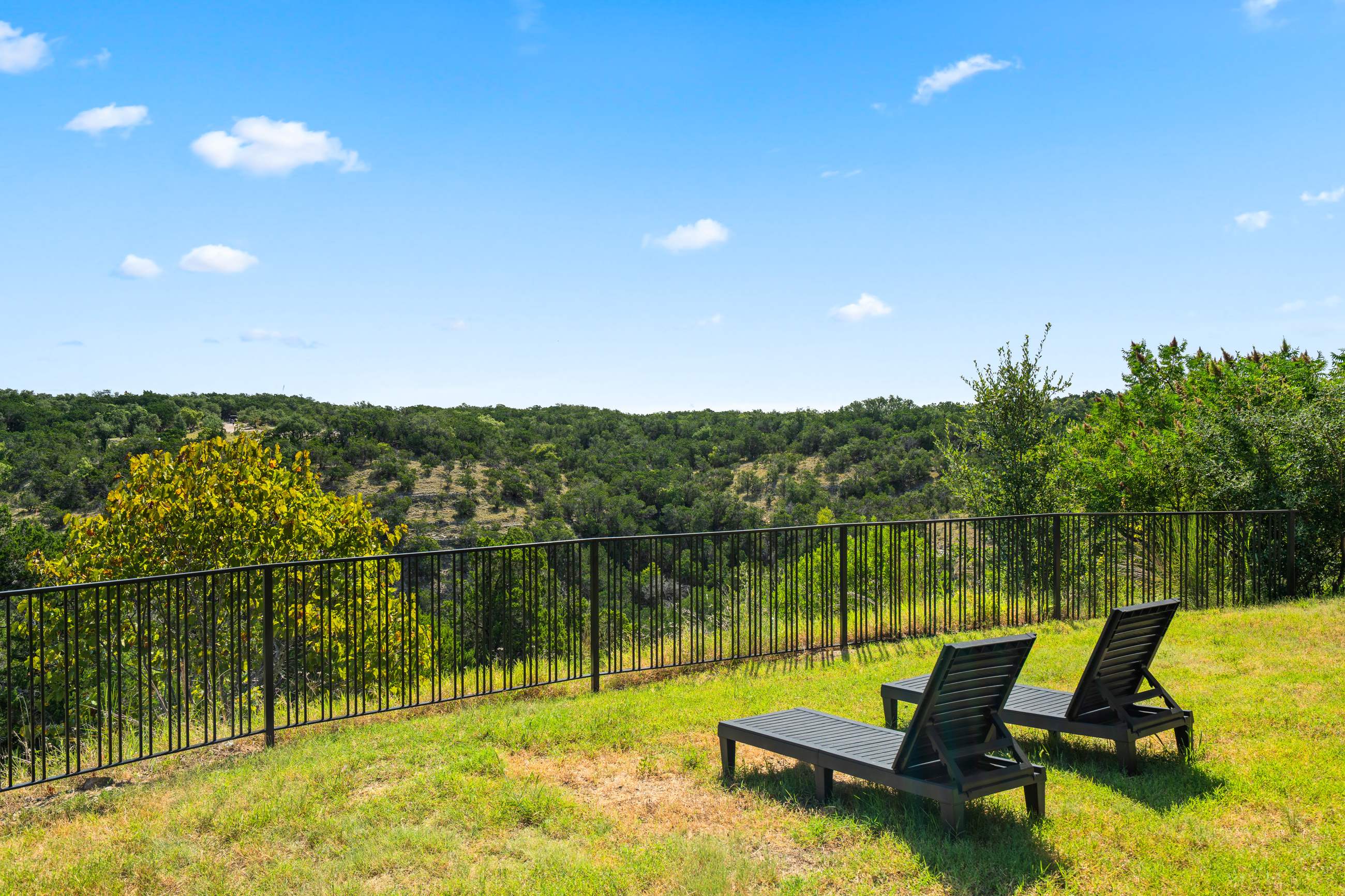Grassy hillside yard with two dark lounge chairs overlooking a vast, tree-covered valley under a clear blue sky. A black metal fence separates the yard from the scenic natural landscape.