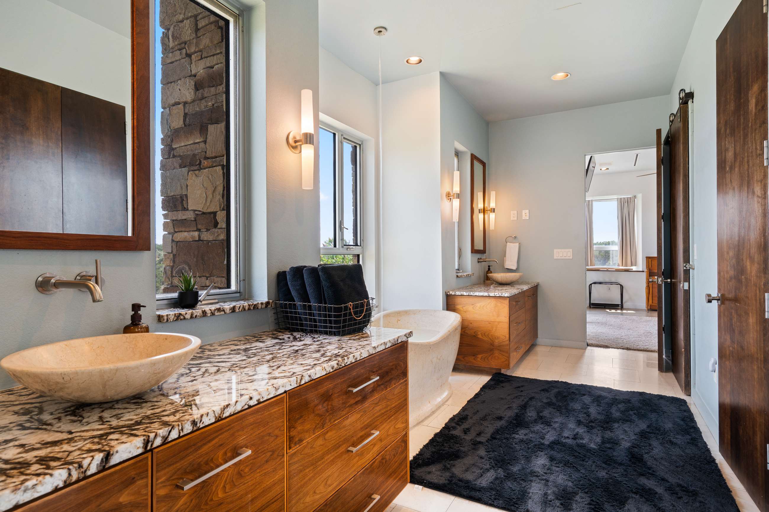 Wide view of a luxurious master bathroom with light blue walls, two wooden vanities with granite countertops and vessel sinks, a freestanding stone tub, and large windows overlooking a natural landscape.