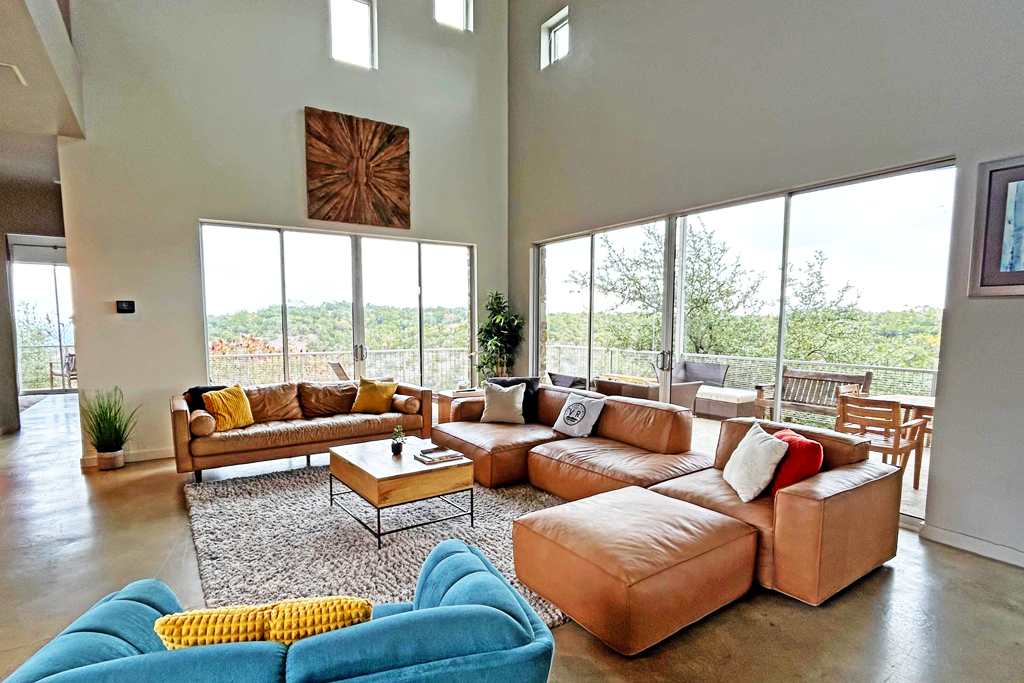 A spacious living room with high ceilings, large floor-to-ceiling windows offering scenic views, and multiple leather sofas arranged around a wooden coffee table on a large rug.