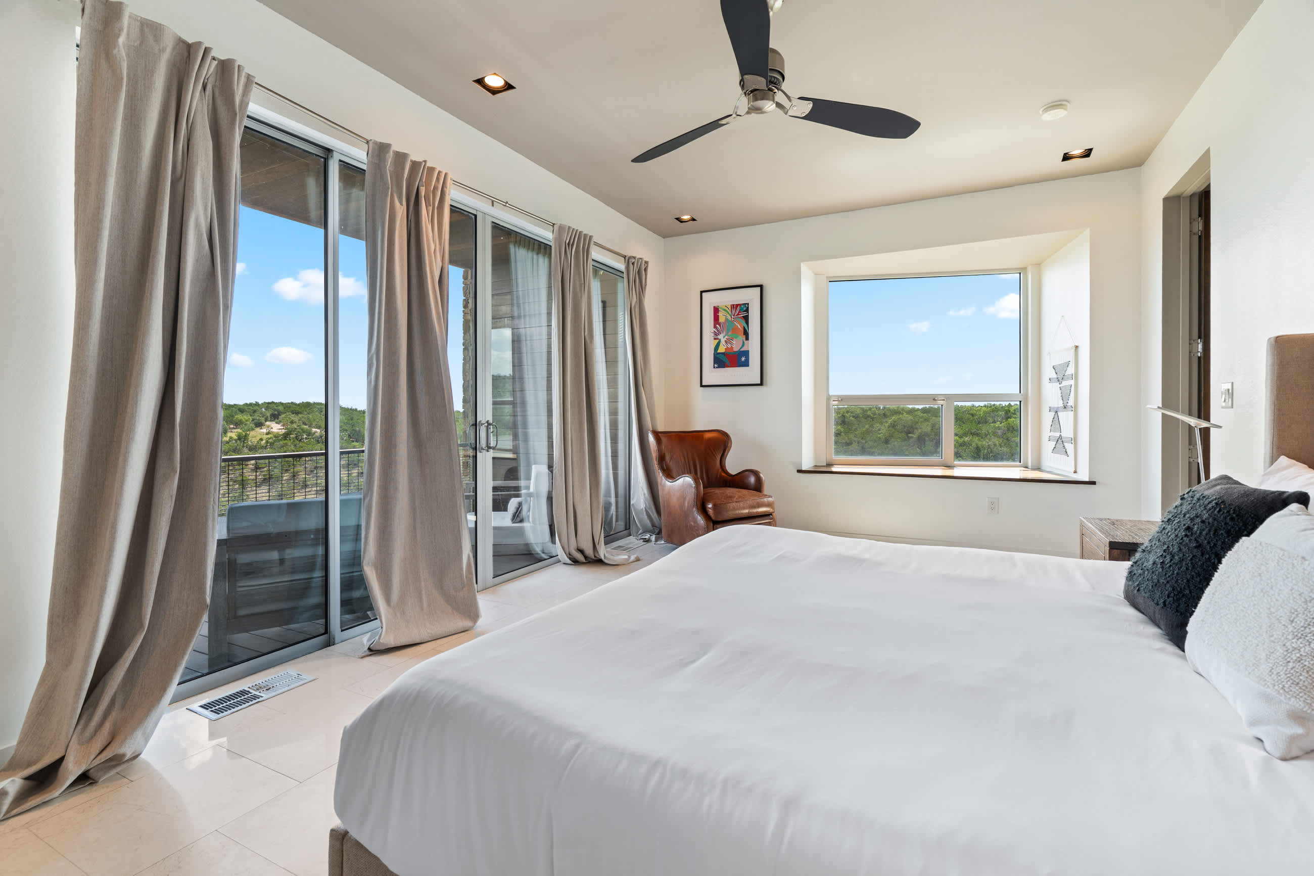 Bright bedroom featuring a white bed, a brown leather armchair, and a large window offering expansive outdoor views. A colorful abstract artwork hangs on the wall.