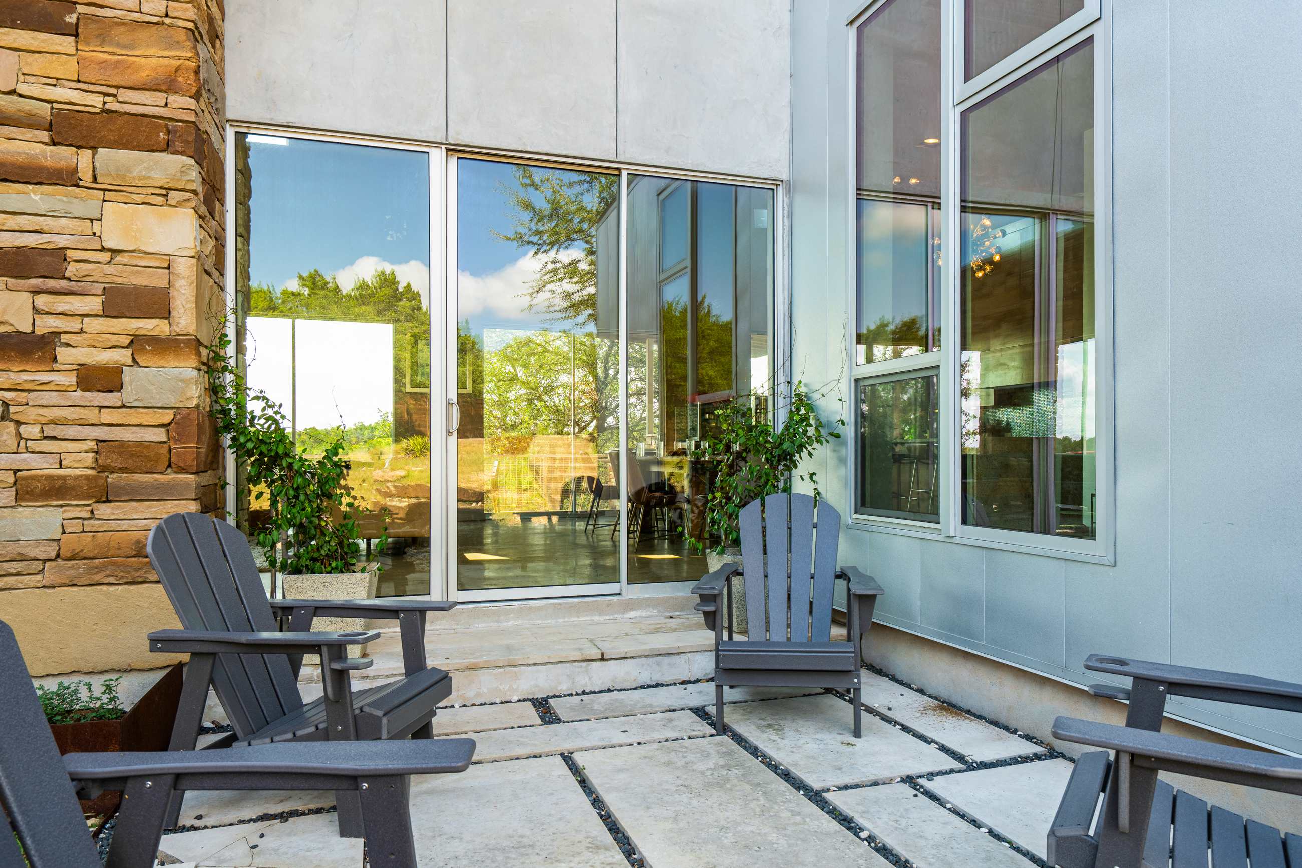 An outdoor seating area with concrete pavers and dark Adirondack chairs, set against a modern building facade with large glass sliding doors and windows, surrounded by natural landscaping.