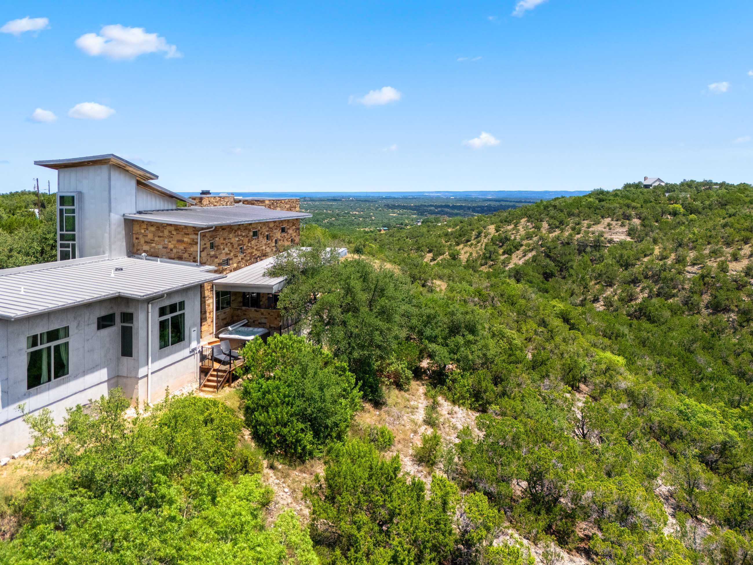 A high aerial shot reveals the contemporary house with its stone and metal elements, blending into the expansive, rugged green hills and distant horizon under a bright sky, highlighting the remote setting.