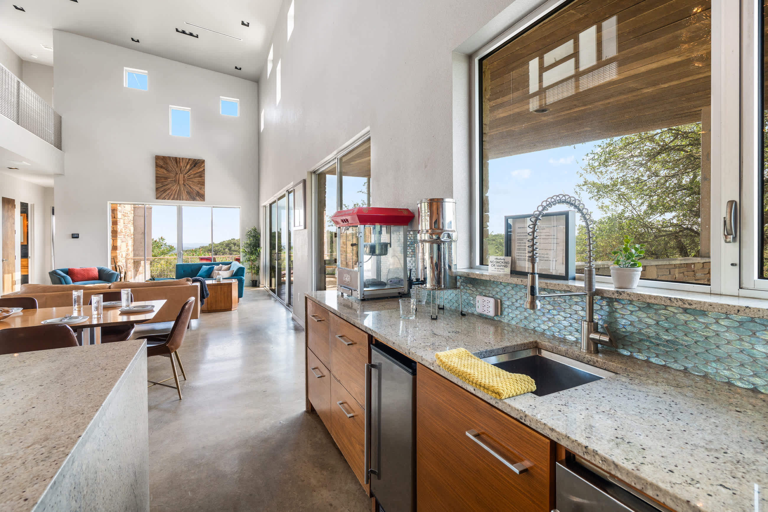 Modern kitchen with light granite countertops, a large window overlooking a natural landscape, and a red popcorn machine, open to a spacious living area with high ceilings.