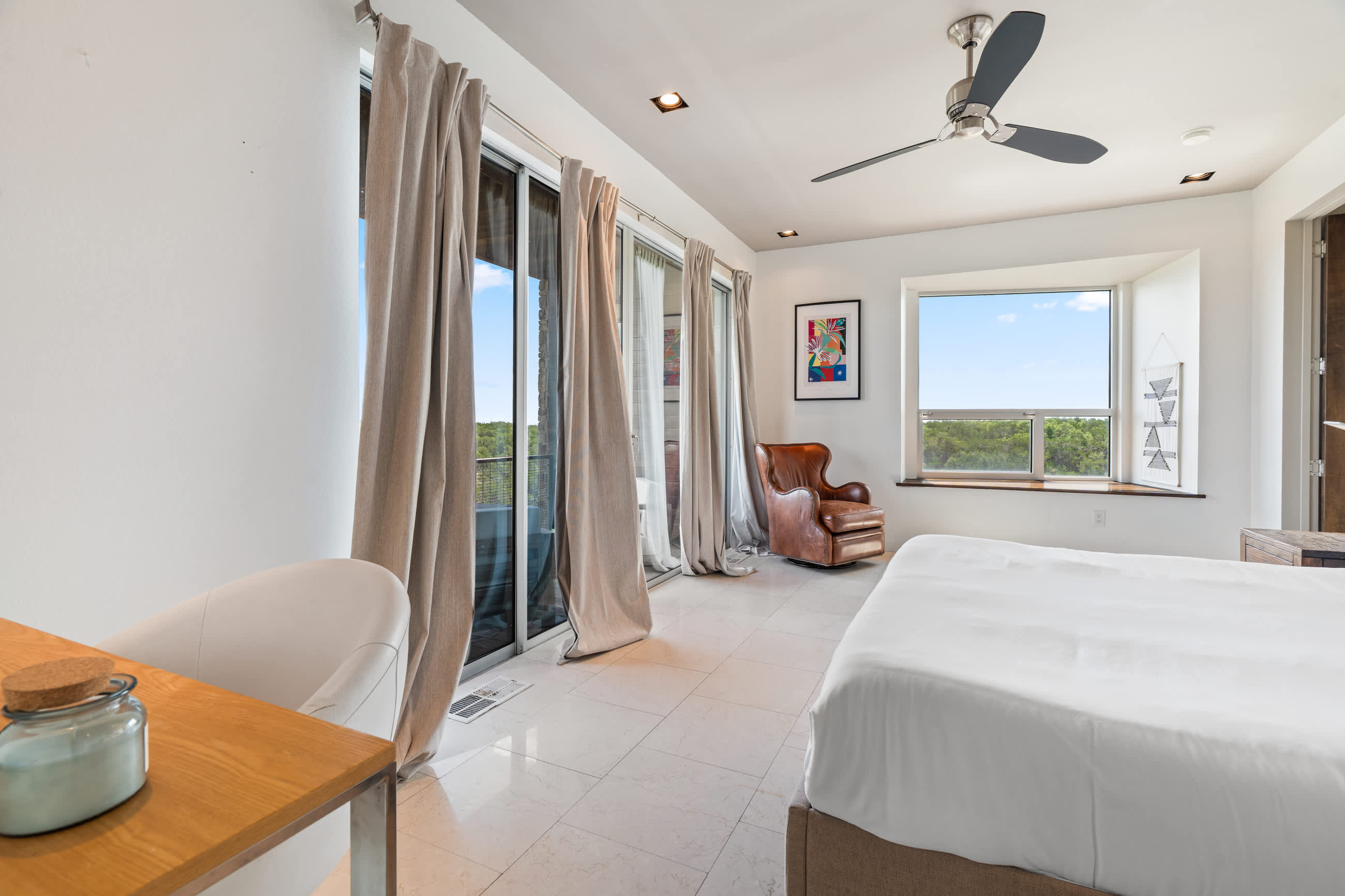 Spacious bedroom with a white bed, a small desk with a white chair, and multiple sliding glass doors opening to a balcony with outdoor views. A ceiling fan is overhead.