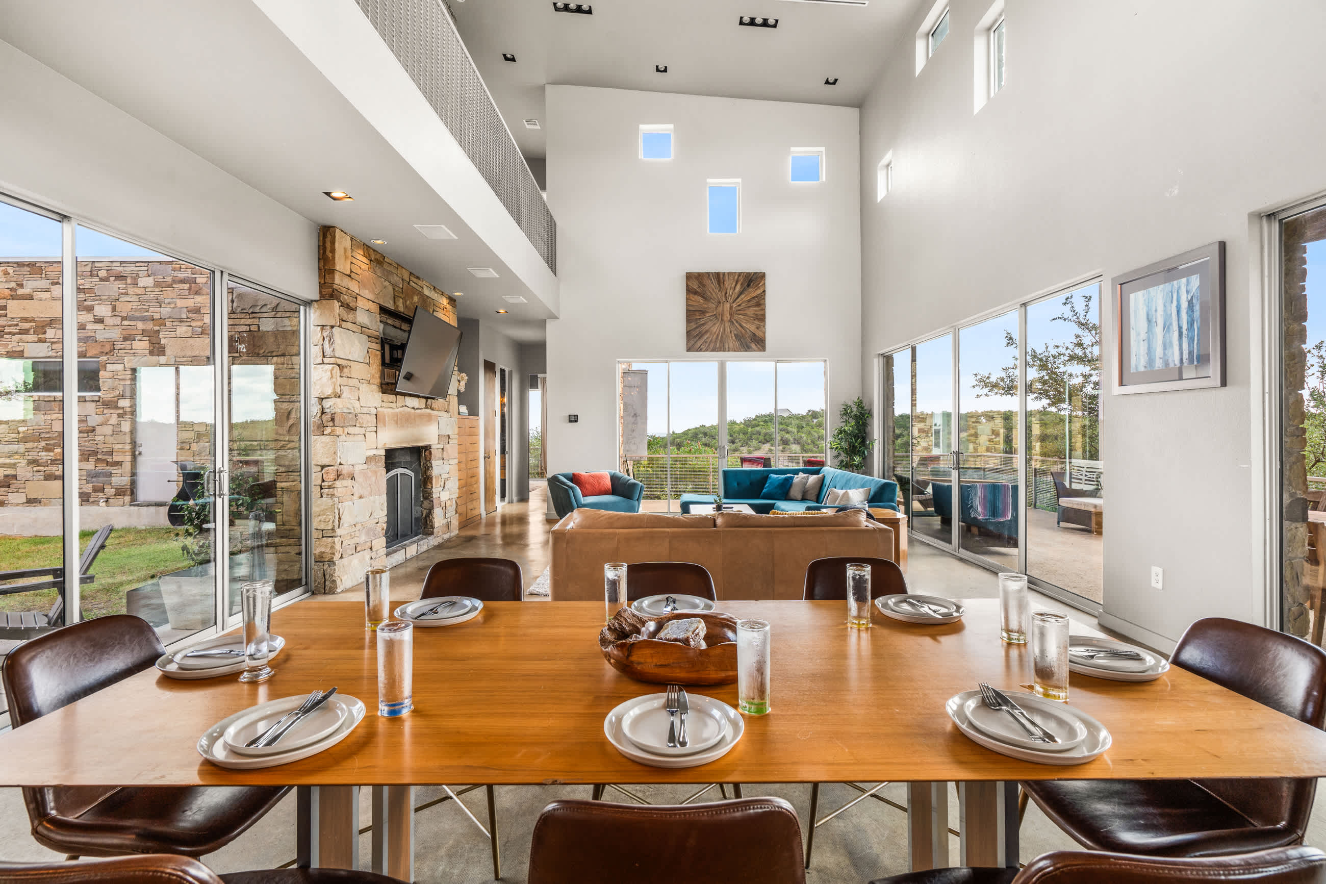 Spacious open-plan living area with a long wooden dining table, a cozy living room with sofas, and a stone fireplace, all bathed in natural light from large windows.