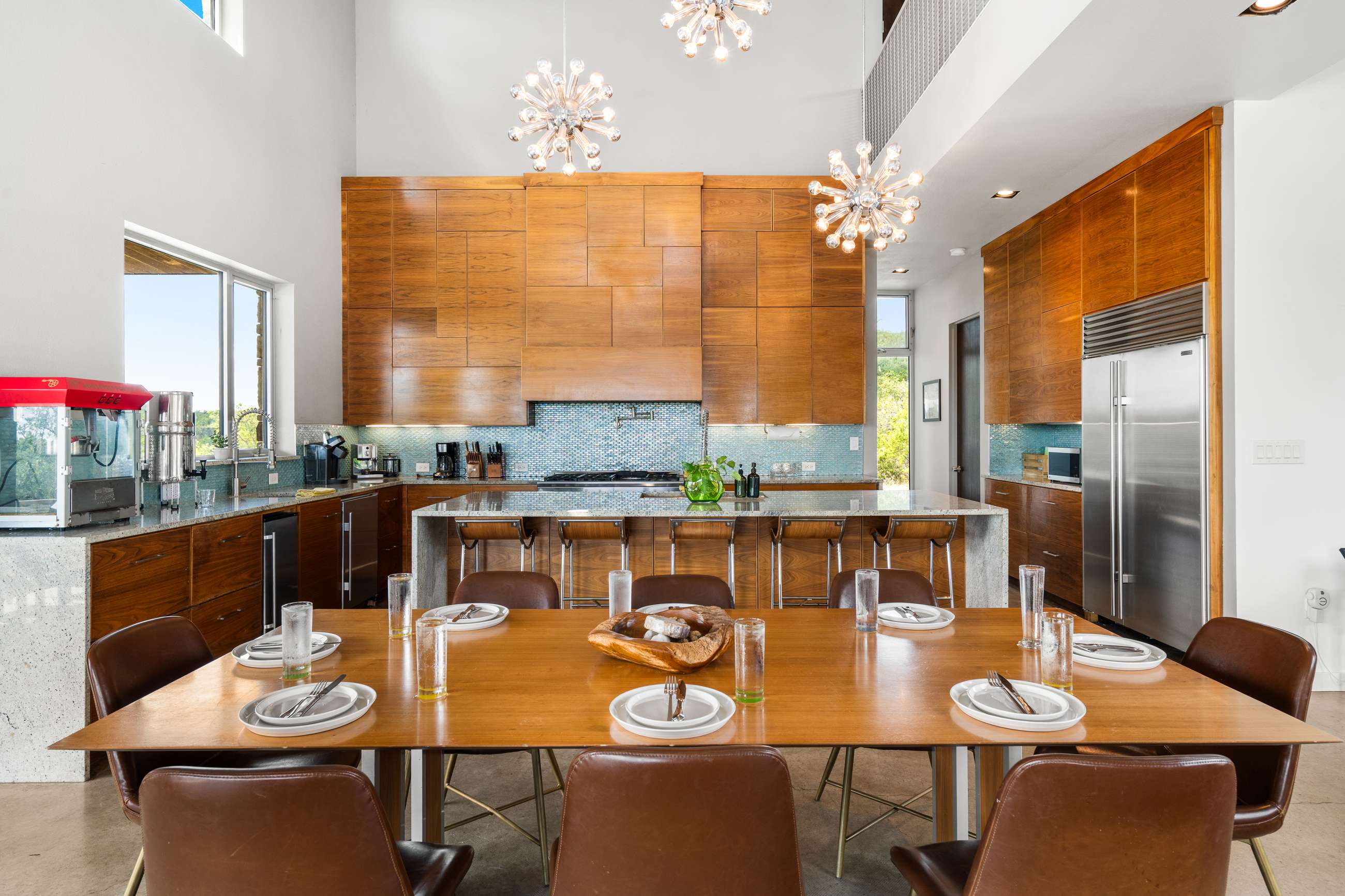 A modern open-concept dining area featuring a long wooden table with brown leather chairs, adjacent to a large kitchen island with bar stools. The space is brightly lit by sputnik chandeliers and large windows, showcasing sleek wooden cabinetry.