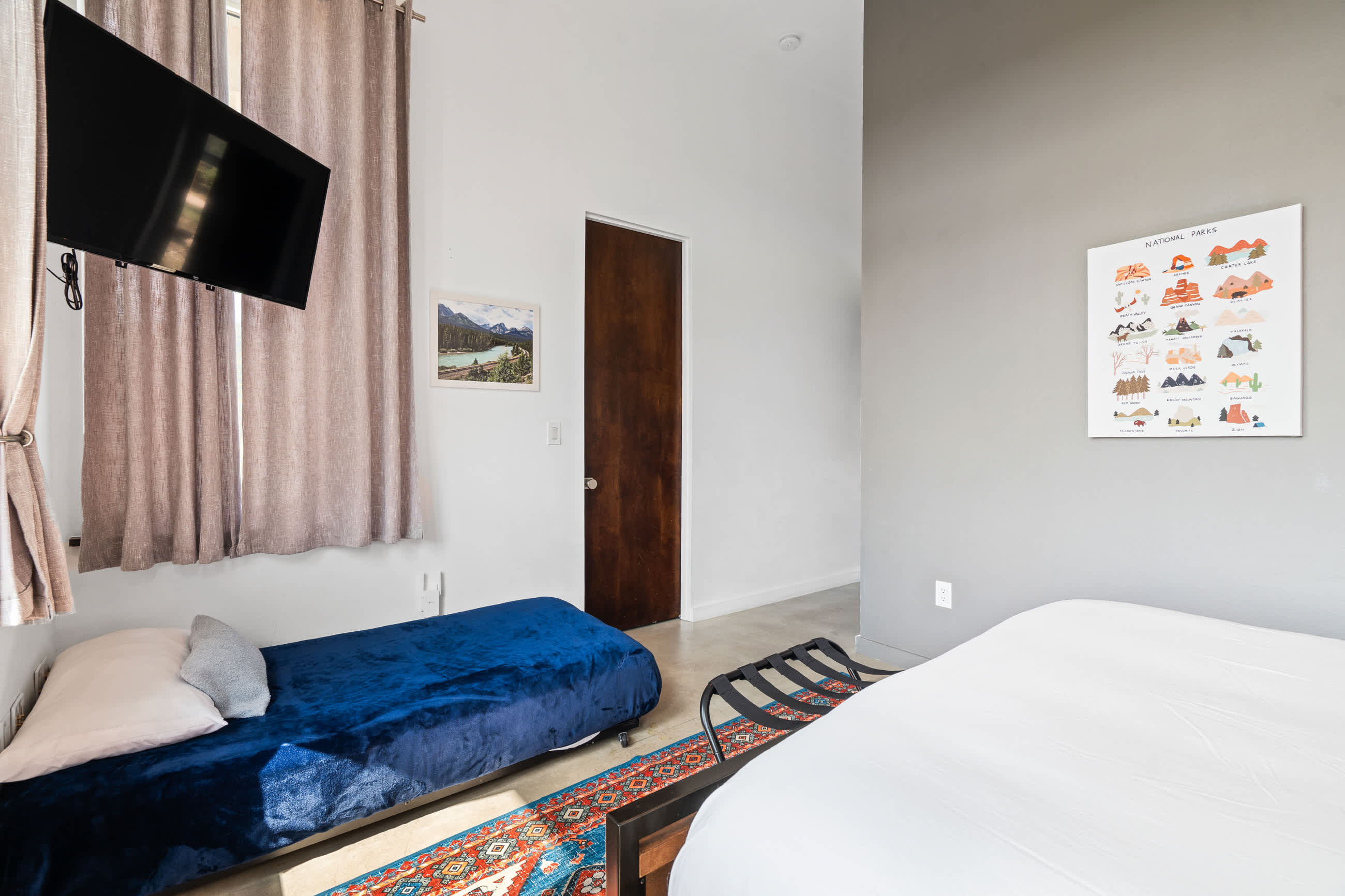 A bright bedroom features a queen-sized bed, a blue velvet twin daybed, and a colorful patterned rug on a concrete floor. A dark wooden door is visible, and a wall-mounted TV hangs near a window.