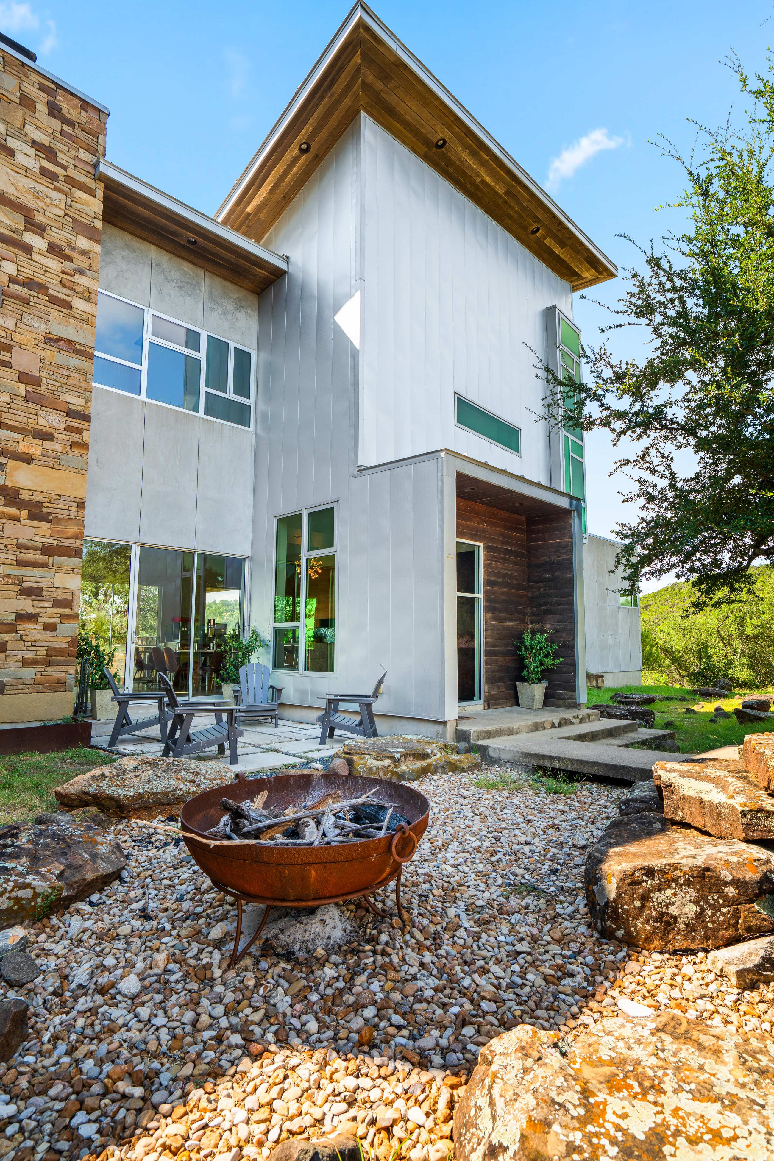 Modern multi-level house exterior with light gray and metal siding, a large stone chimney, and a wooden entrance. A rustic fire pit sits on a gravel patio with large natural rock landscaping and outdoor chairs.