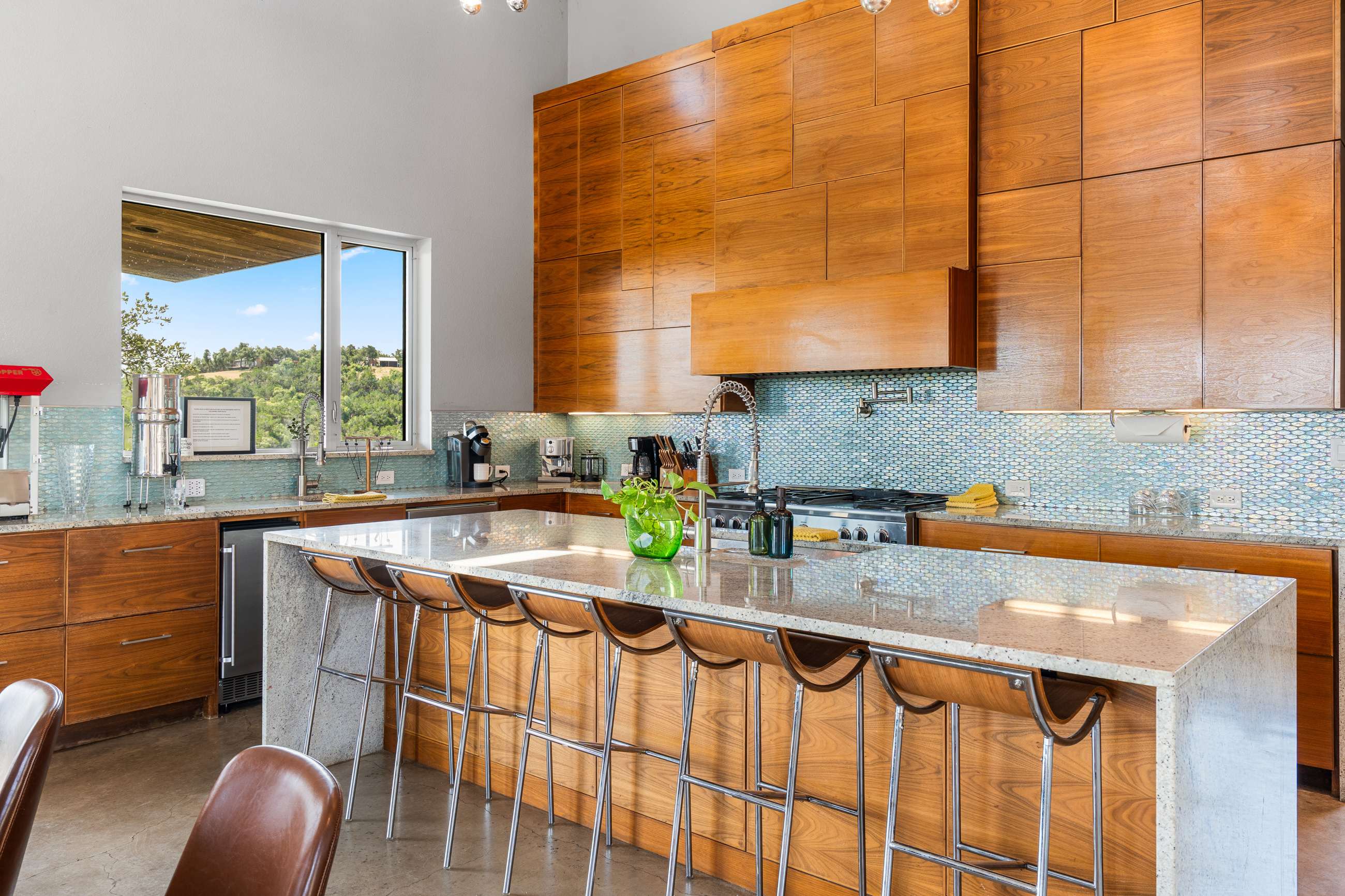 Spacious modern kitchen with sleek wooden cabinetry, light granite countertops, and a blue mosaic tile backsplash. A large island with bar stools provides ample seating, and a window offers scenic outdoor views.