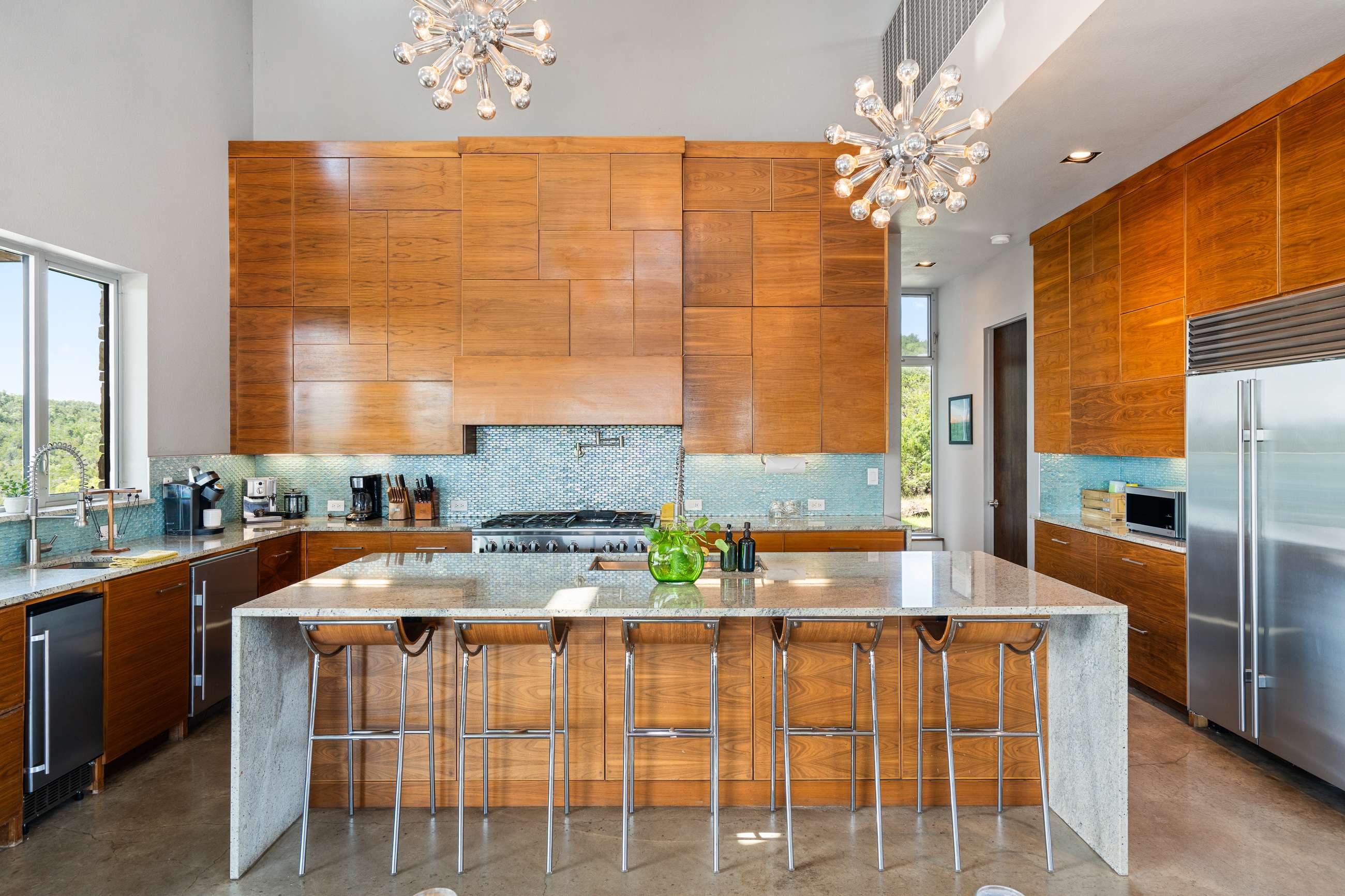 Bright, high-ceilinged kitchen featuring extensive wooden cabinetry, a large granite island with bar seating, and a vibrant blue mosaic tile backsplash. Two modern sputnik chandeliers illuminate the space, complementing the stainless steel appliances.