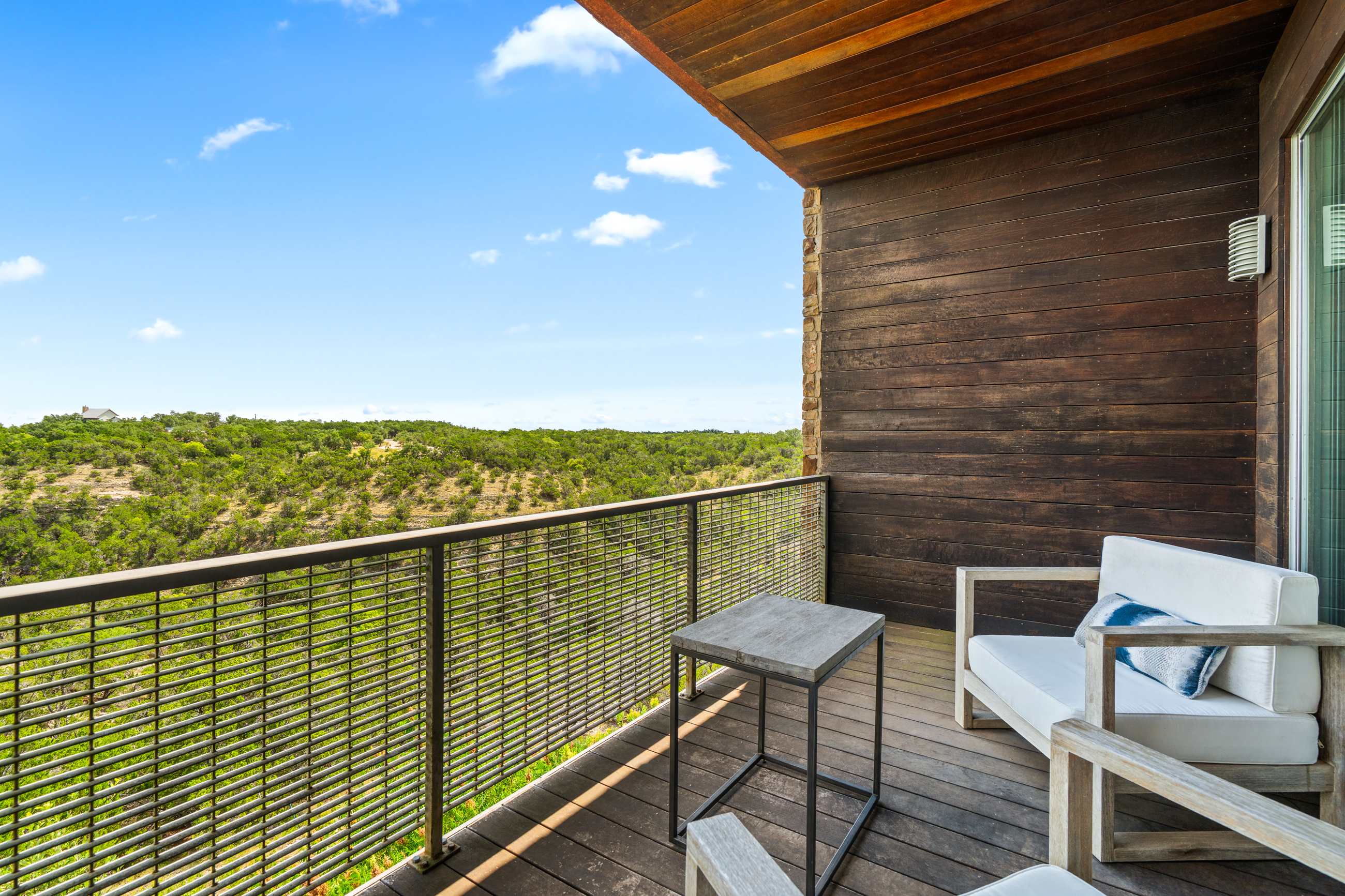Modern balcony with wooden decking and a metal railing, featuring two white outdoor chairs and a small table, set against a backdrop of lush green hills and a clear blue sky. The building exterior is visible.