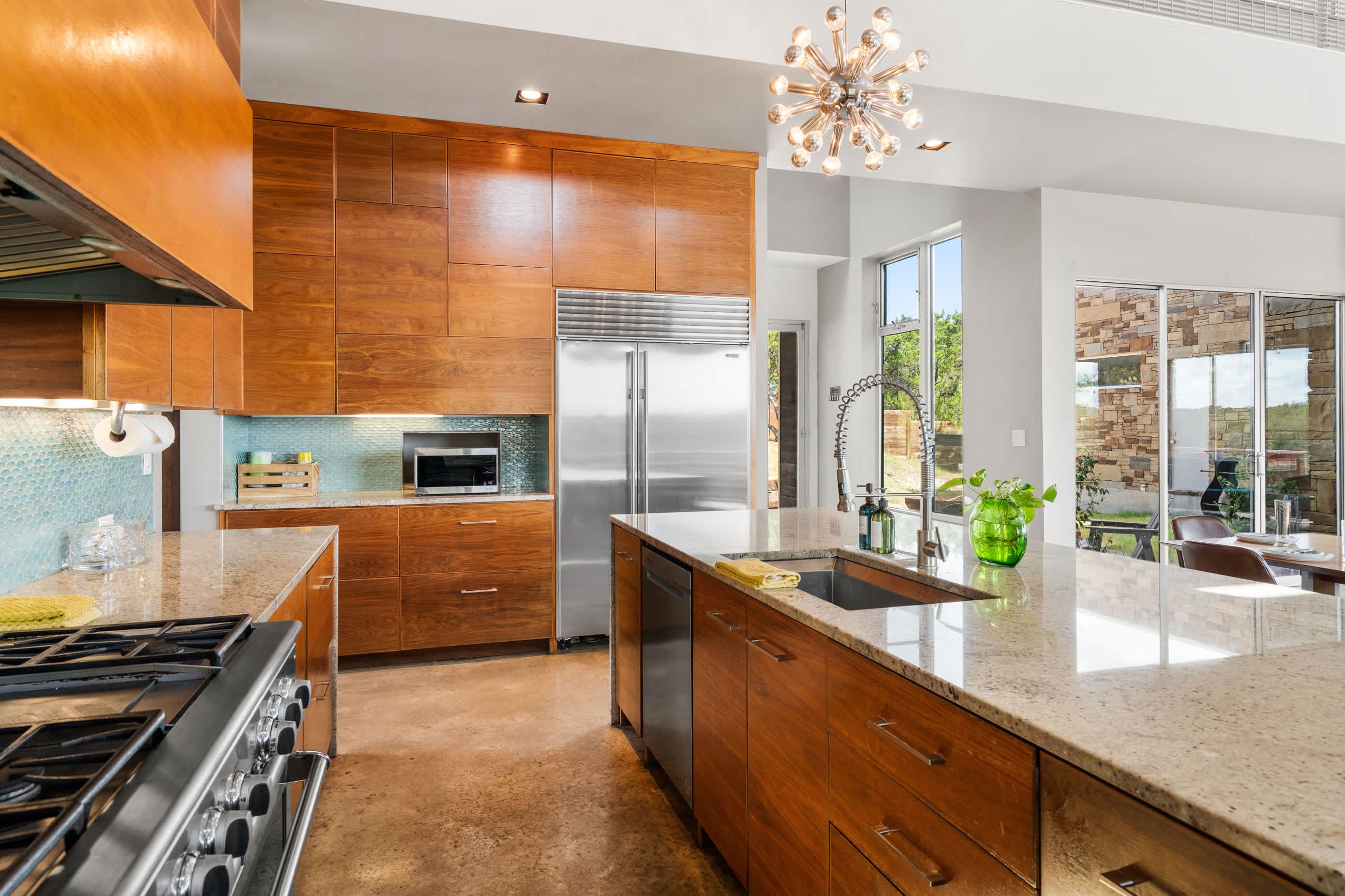 A wide shot of the modern kitchen, highlighting the large stainless steel refrigerator, extensive wooden cabinetry, and a granite island with a sink. The room features high ceilings, a sputnik chandelier, and large windows.