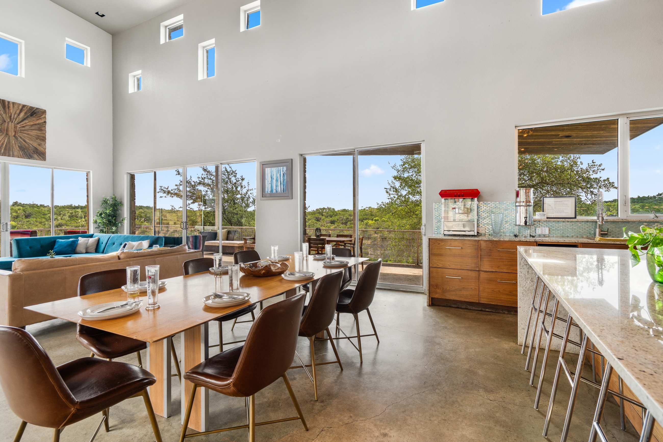 An expansive open-concept living space featuring a long wooden dining table in the foreground and a comfortable brown leather sectional sofa in the background. Large floor-to-ceiling windows offer panoramic views of the natural landscape.