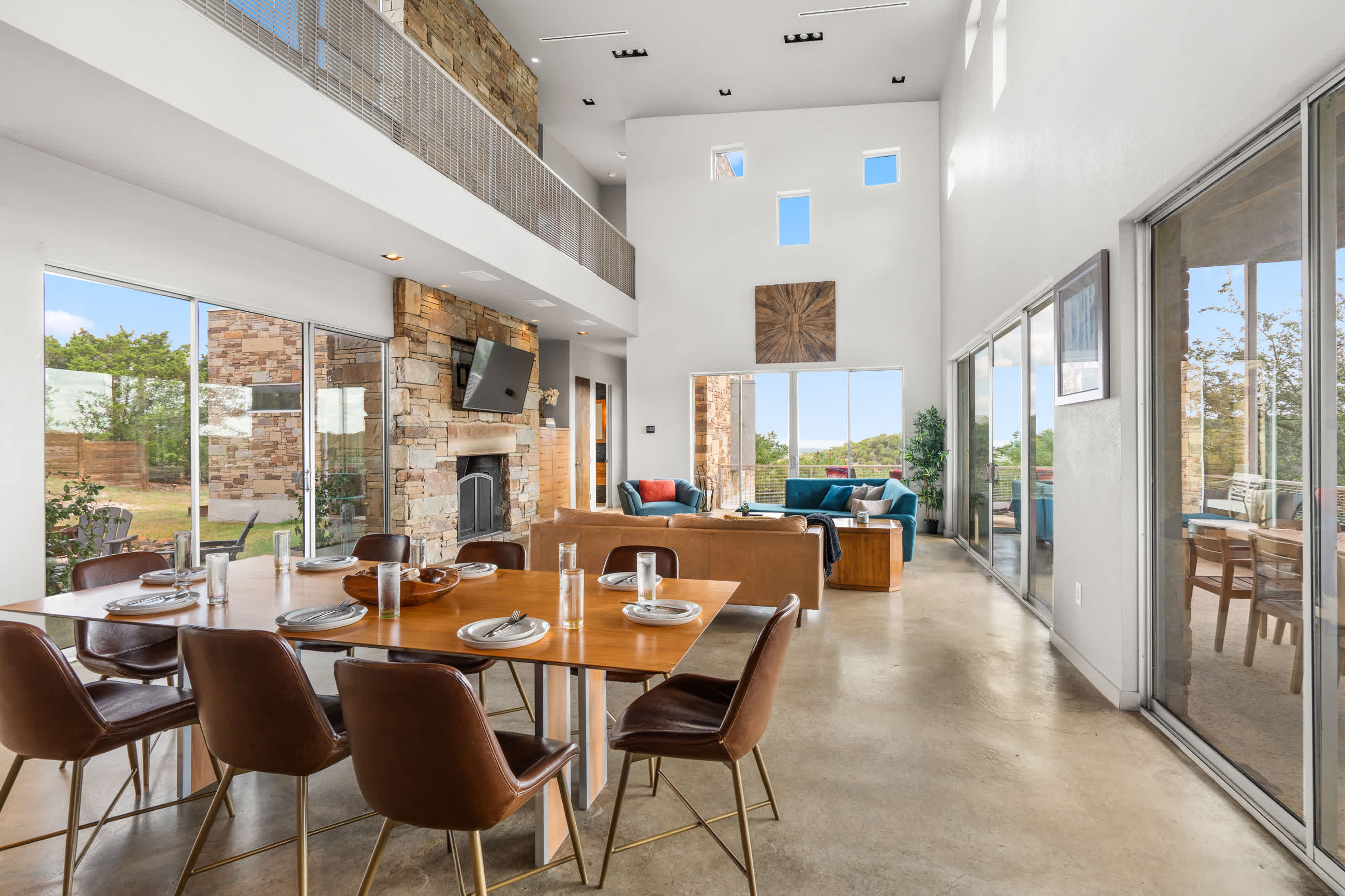 Grand open-concept living space featuring a long dining table, a comfortable living area, a prominent stone fireplace, and an upper-level balcony overlooking the main floor.