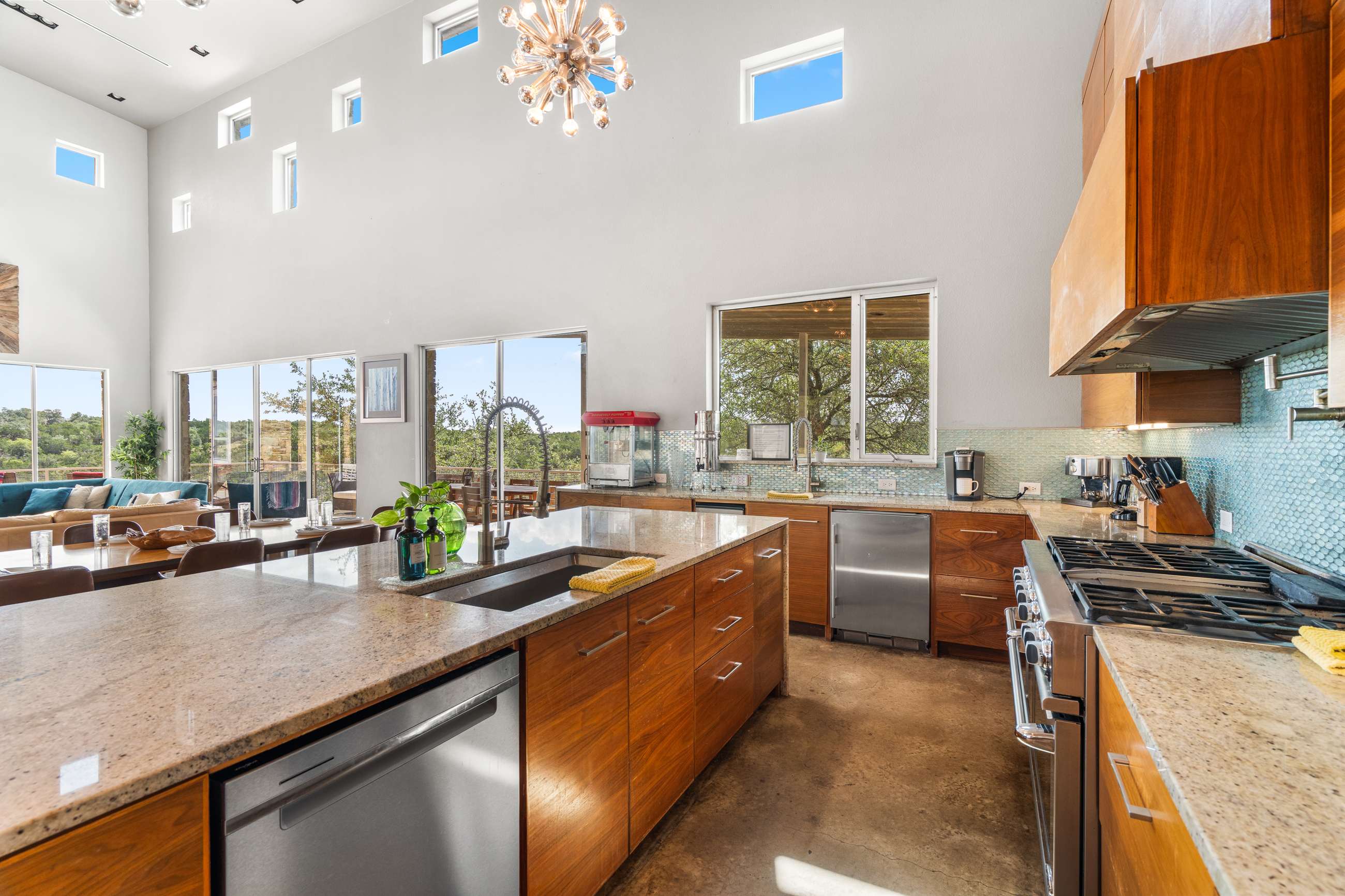 An overhead view of the spacious kitchen, showing the large granite island with a sink and two dishwashers, facing a stainless steel range and light blue mosaic tile backsplash. The room has high ceilings and multiple windows.