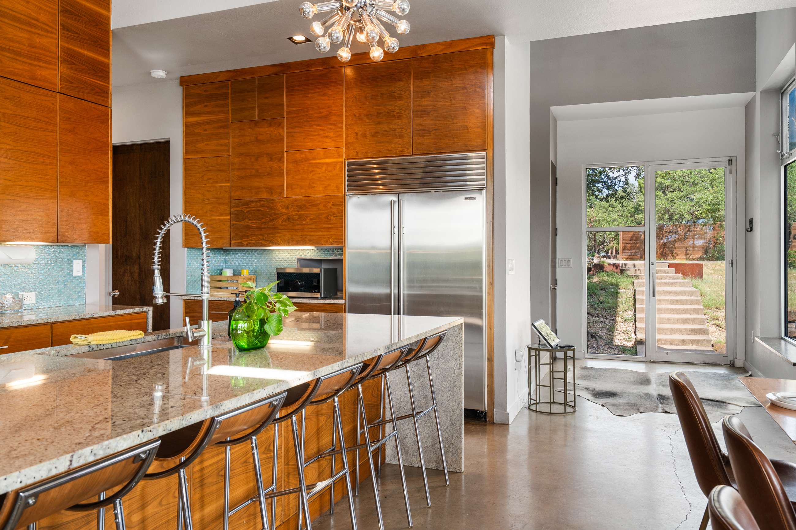 A modern kitchen featuring a large stainless steel refrigerator, extensive wooden cabinetry, and a granite-topped island with a sink and bar seating. The space has concrete floors and a view towards an exterior door and steps.