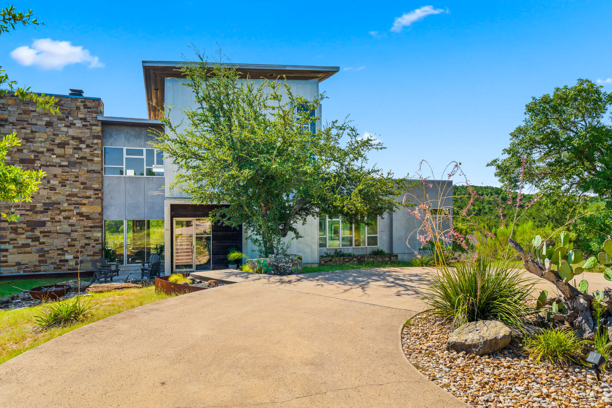 A modern house entrance with a concrete driveway, featuring a blend of stone and light-colored siding, large windows, and natural landscaping including a prominent tree and desert plants.