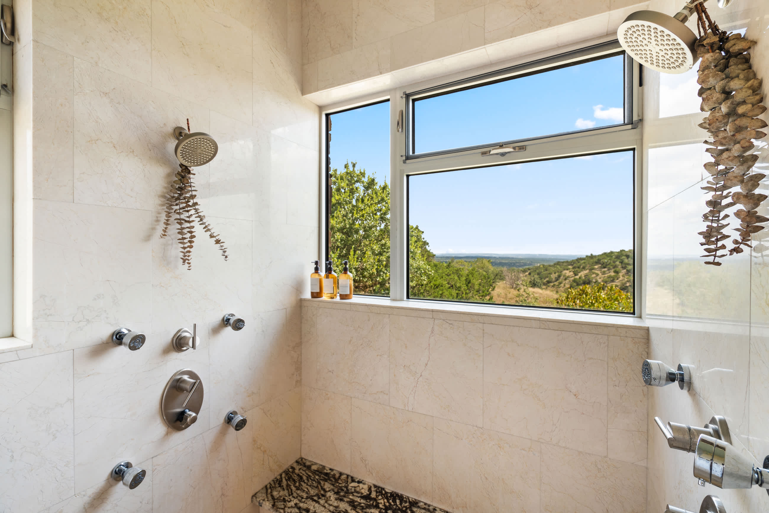 Spacious walk-in shower with light marble-look tiles, multiple shower heads, and a large window offering panoramic views of the outdoors. A granite bench is integrated into the shower.