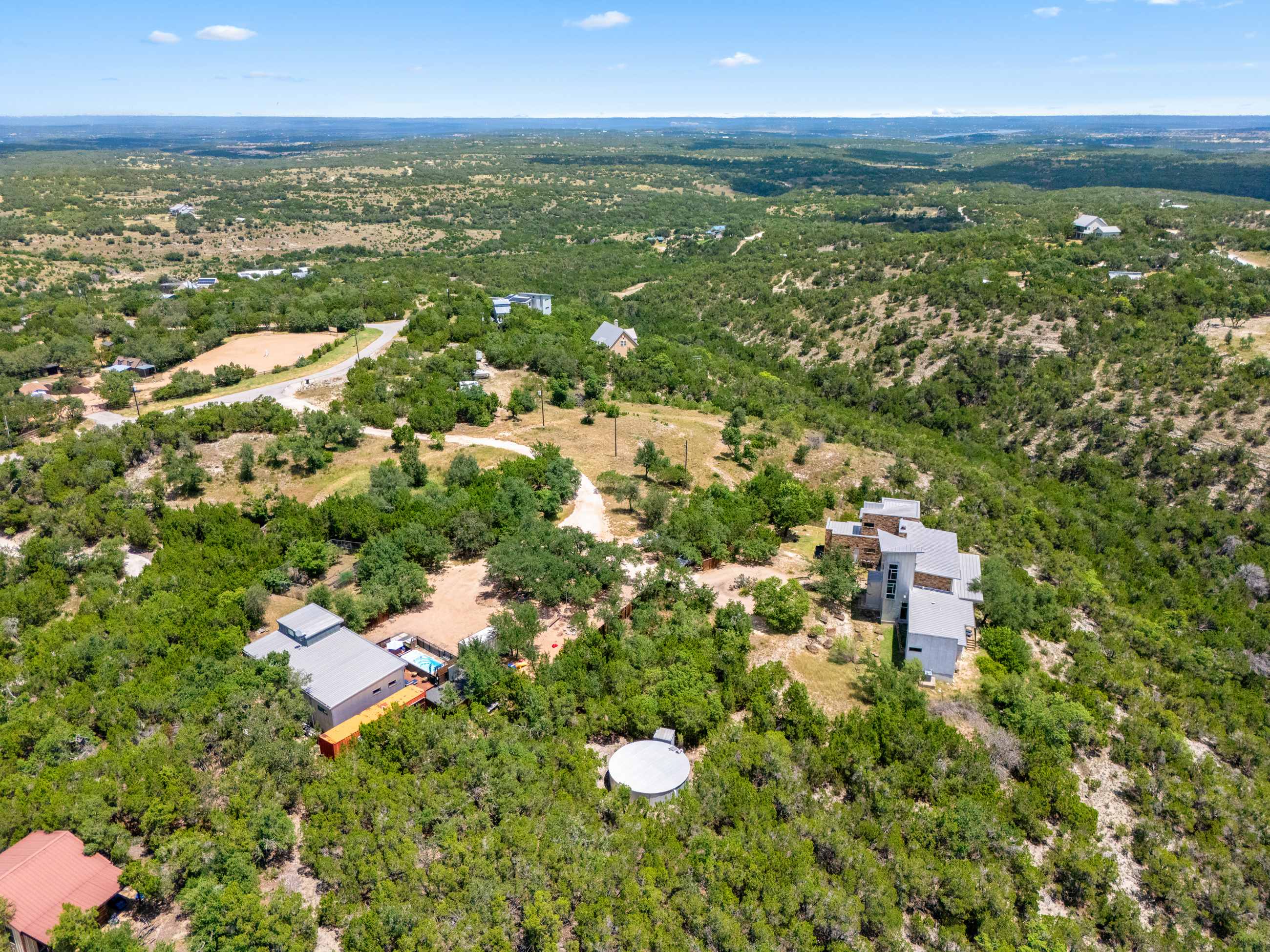 An expansive aerial view showcases a luxury hilltop retreat nestled within a vast, green, and hilly landscape under a clear blue sky. Multiple modern and traditional homes are scattered among dense trees, connected by winding dirt roads, offering panoramic views of the surrounding natural terrain.