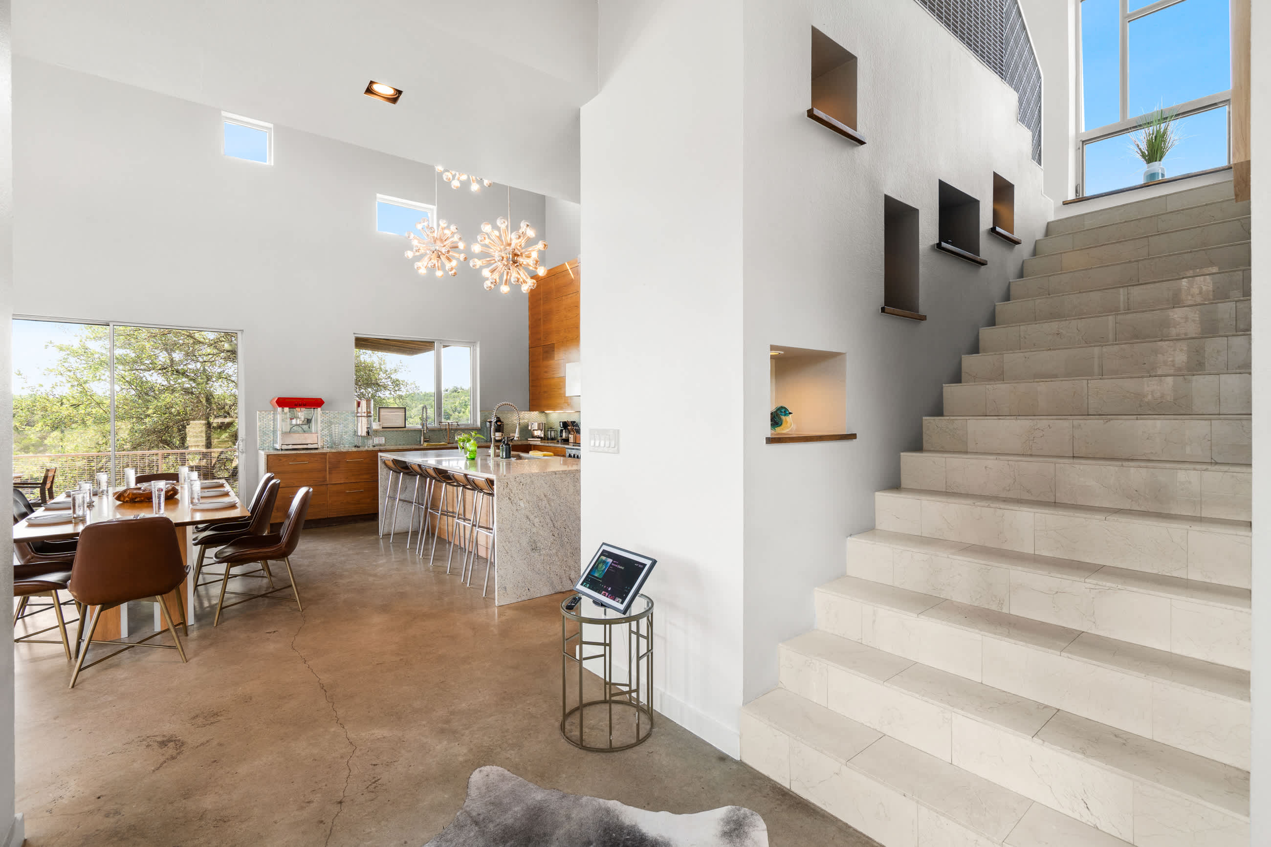 A modern open-concept dining area with a long wooden table and brown leather chairs, concrete floors, and a view of the kitchen with a large island and contemporary light fixtures. A white staircase with built-in shelves is visible on the right.