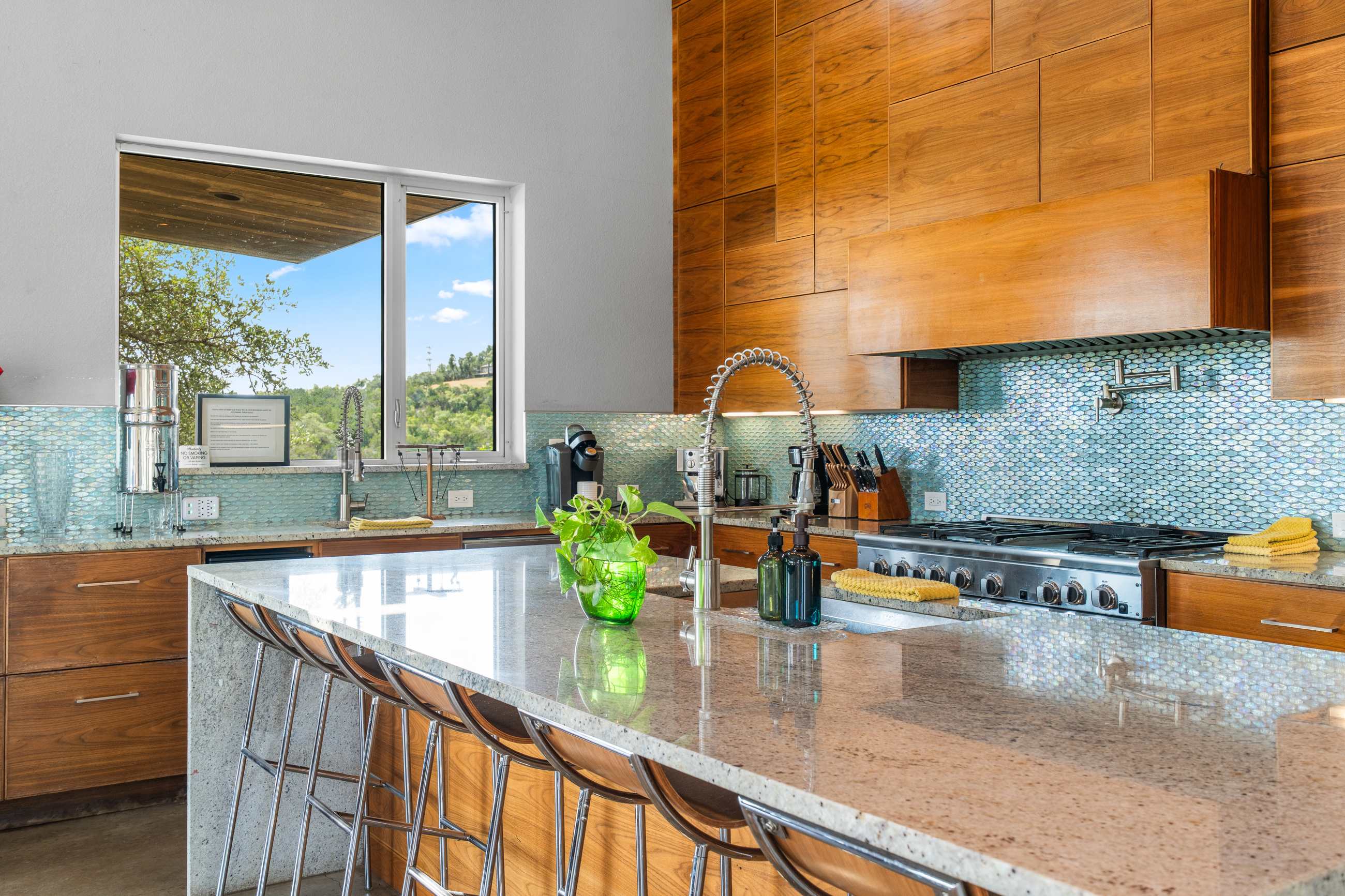 Detailed view of the modern kitchen showing a granite countertop, blue mosaic tile backsplash, and a large window with scenic outdoor views. A gooseneck faucet, a coffee maker, and a plant are visible on the counter.
