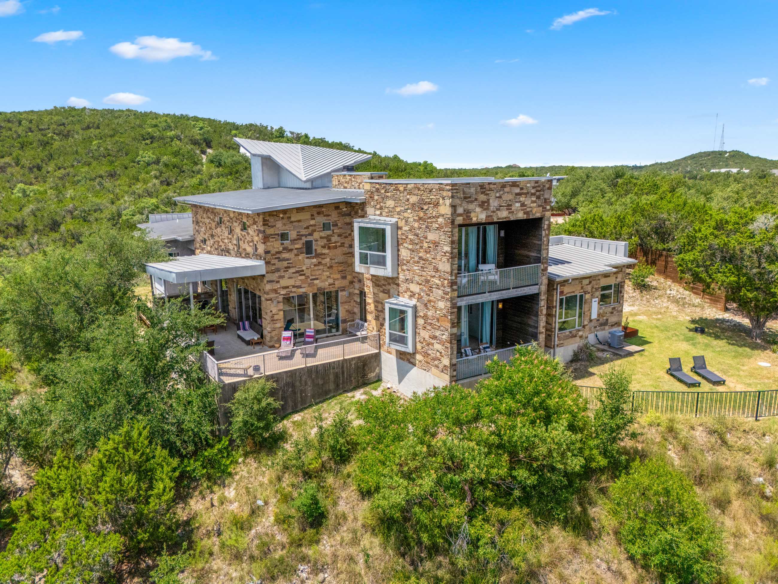 A wide aerial shot displays the contemporary stone and metal house with multiple levels and large windows, surrounded by lush green trees and a manicured lawn area, set against a hilly backdrop.