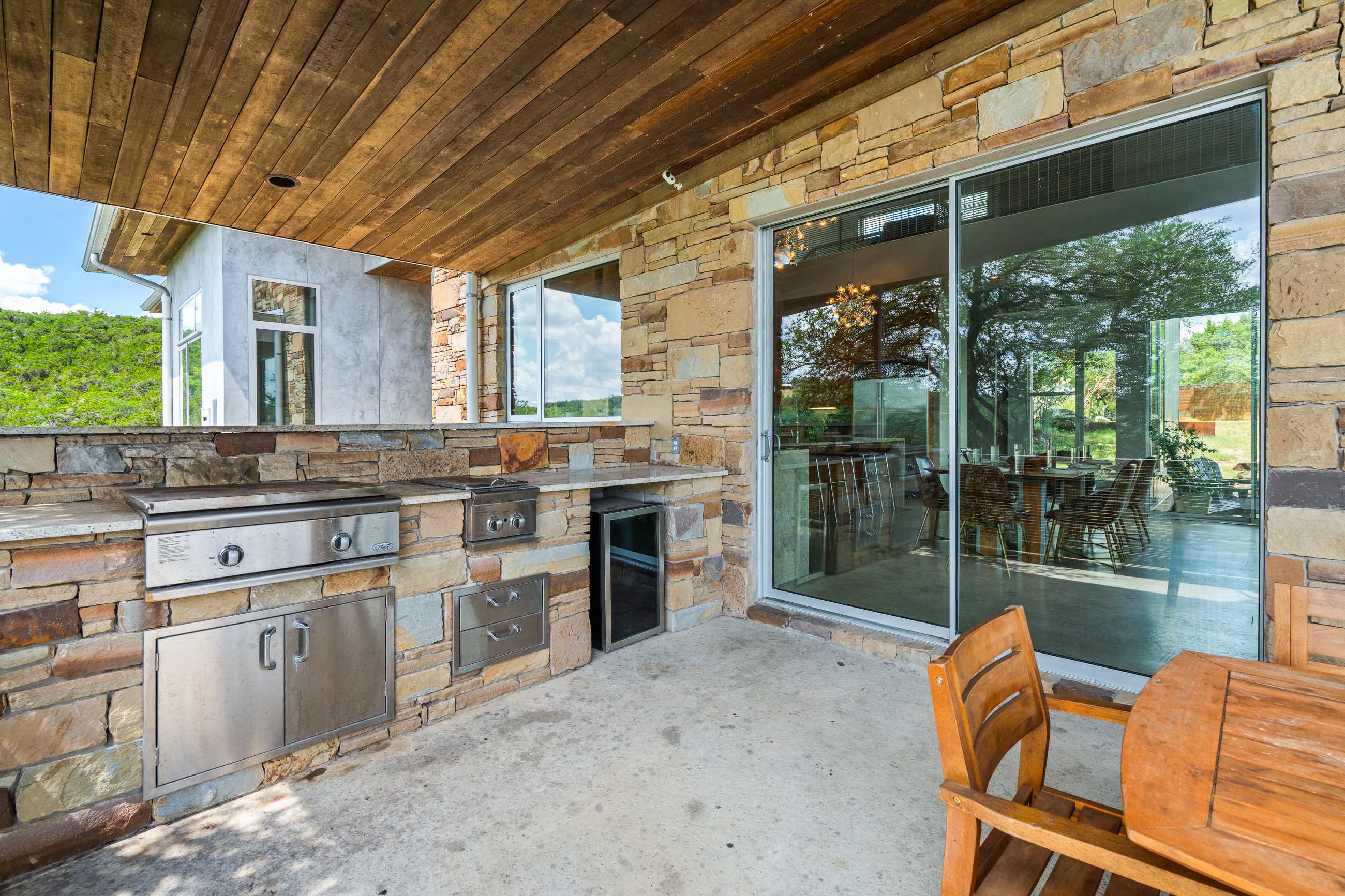 An outdoor kitchen built into a stone wall features a stainless steel grill, a sink, and a mini-fridge, situated on a concrete patio next to large sliding glass doors leading indoors.