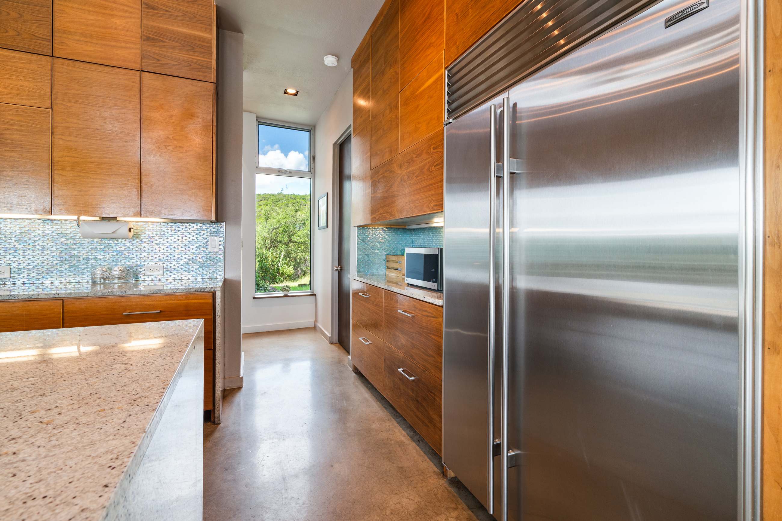 Sleek kitchen hallway featuring warm wooden cabinetry, a large stainless steel refrigerator, and a microwave, with a narrow window offering a glimpse of greenery.