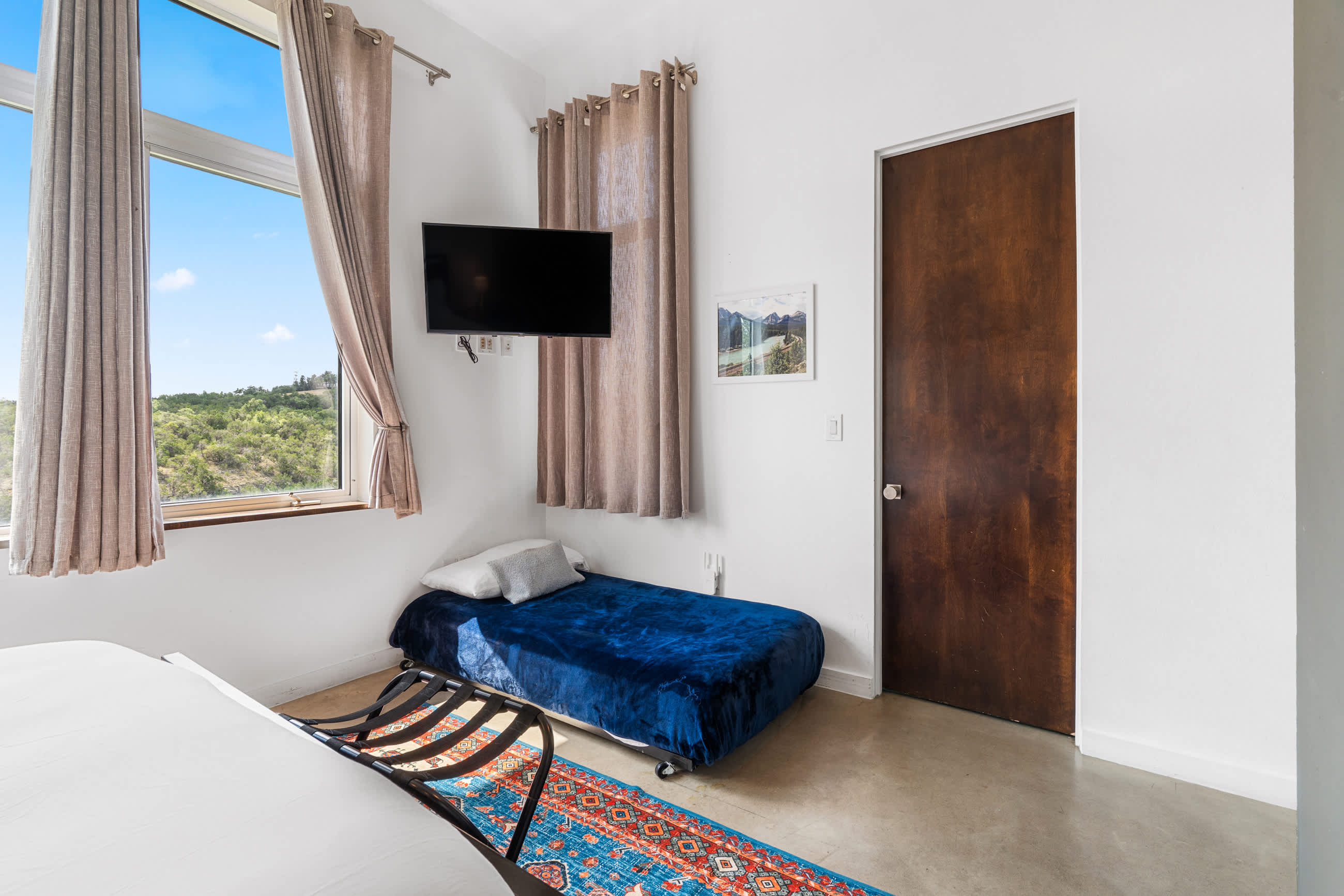 This bedroom includes a queen-sized bed, a blue velvet twin daybed with pillows, and a wall-mounted TV. A colorful patterned rug partially covers the concrete floor, and a window provides natural light.
