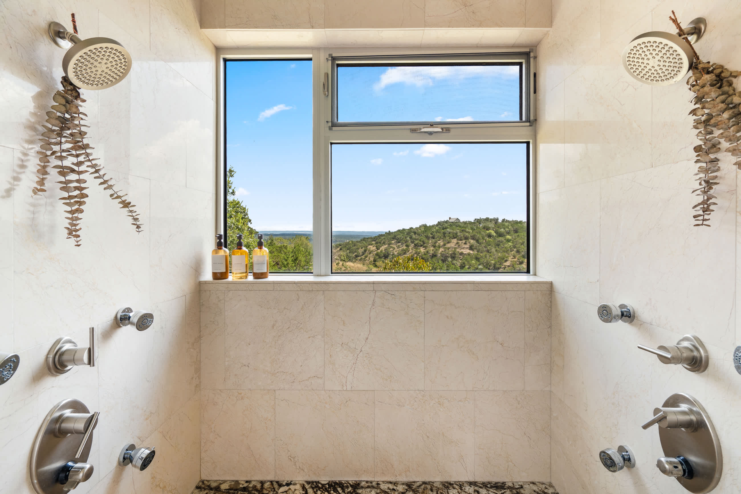 Expansive walk-in shower featuring light marble-look tiles, dual rain shower heads, multiple body jets, and a large window providing stunning views of the surrounding hills and sky.
