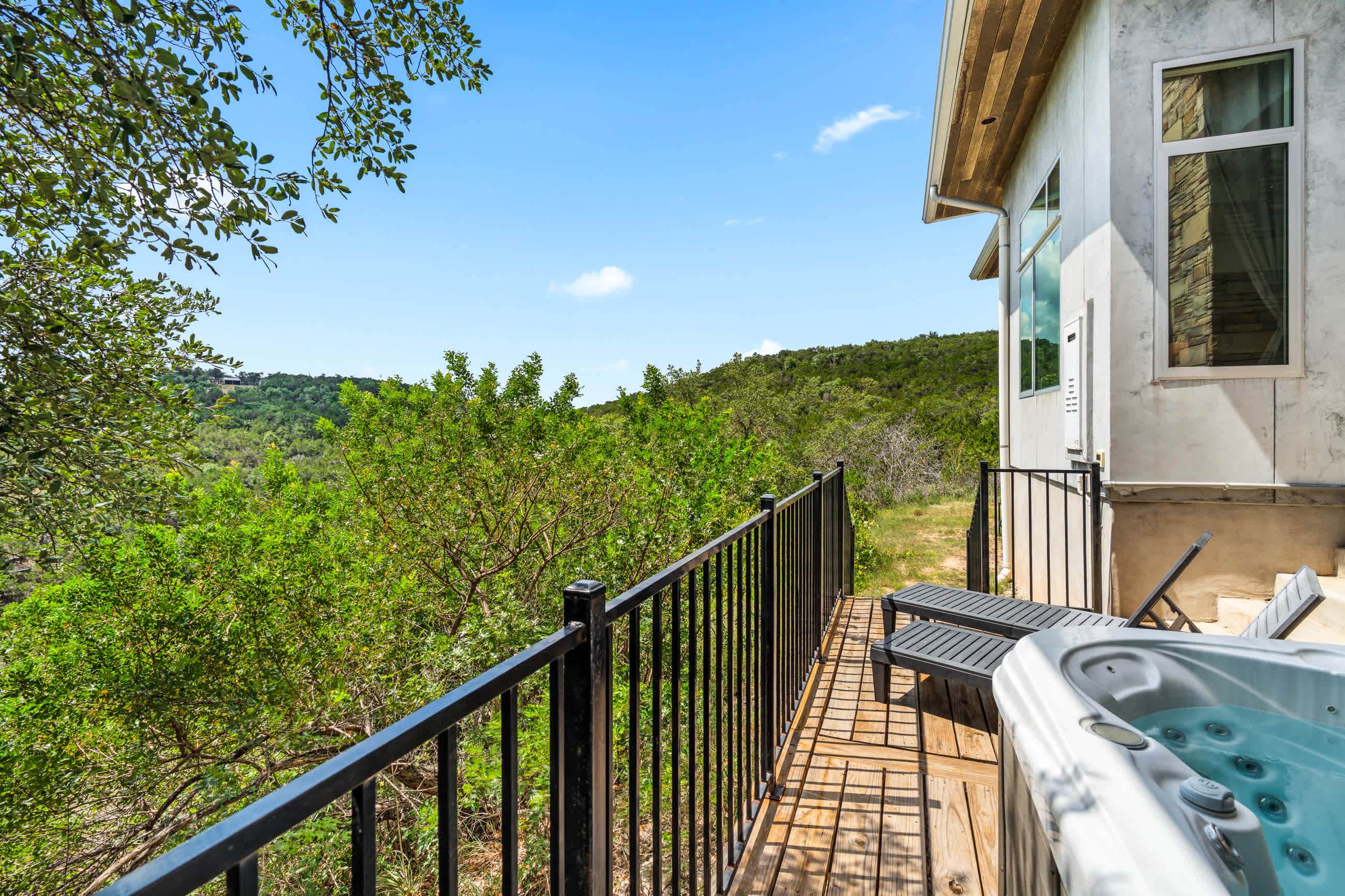 Outdoor hot tub situated on a wooden deck with a black metal railing, offering a private setting with lush green trees and distant hills as a backdrop. Two lounge chairs are nearby.