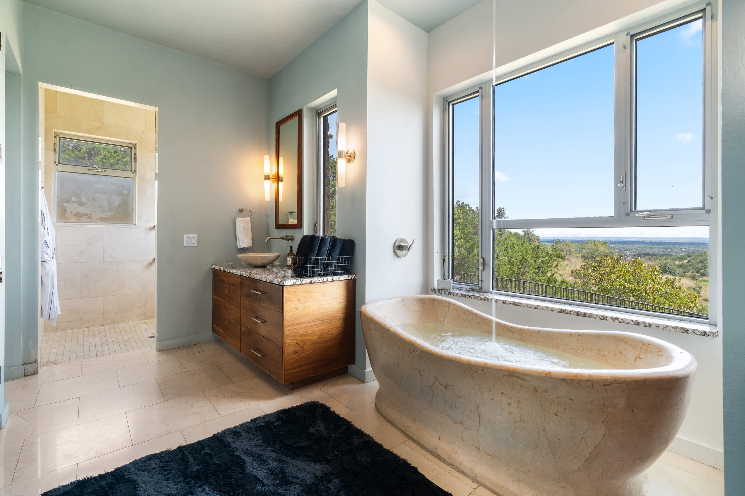 A luxurious bathroom with a freestanding stone bathtub positioned by a large window offering scenic views, a modern vanity with a vessel sink, and a separate walk-in shower visible in the background.