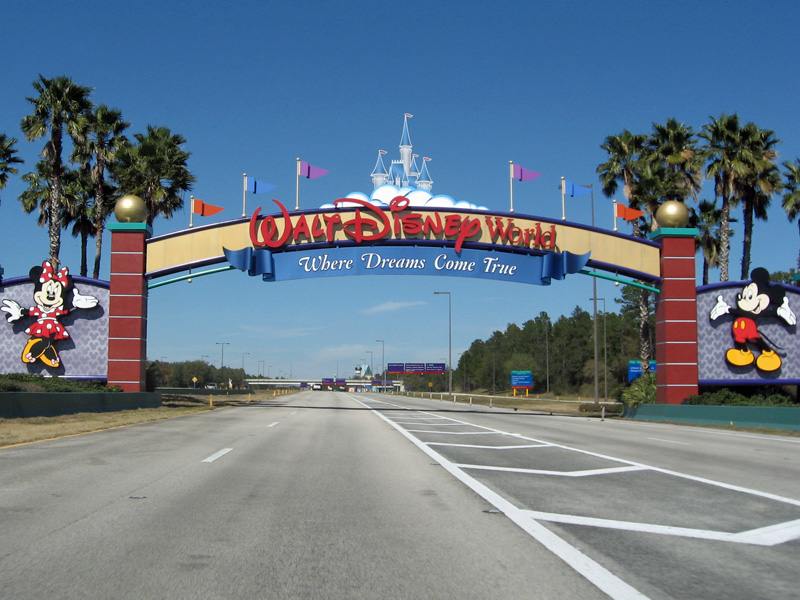 The iconic Walt Disney World entrance archway spans a multi-lane road under a clear blue sky, featuring Mickey and Minnie Mouse characters on the pillars and a castle silhouette above the 'Where Dreams Come True' banner, flanked by palm trees.