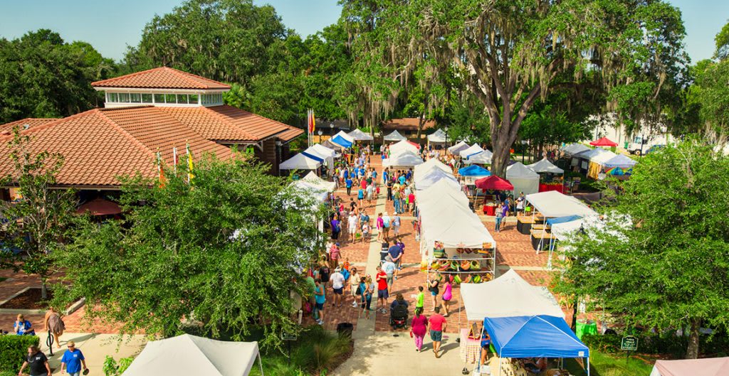 Lively aerial view of an outdoor market or festival with numerous white tents, bustling crowds, and mature trees providing shade, set in a town square with a building featuring a red tile roof.