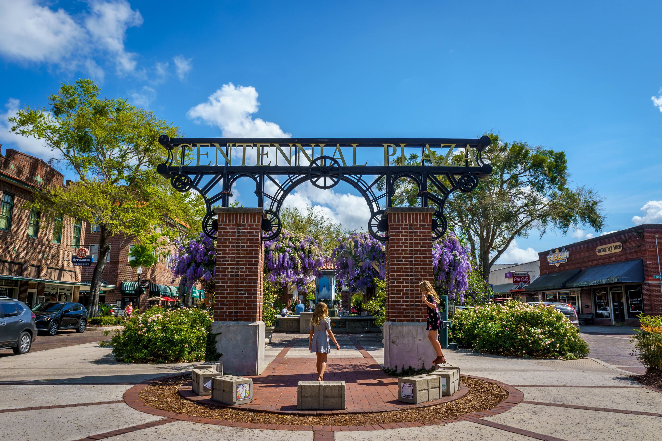 Iconic "Centennial Plaza" archway made of wrought iron and brick pillars, surrounded by lush green bushes and vibrant purple wisteria, with historic buildings and a clear blue sky in the background.