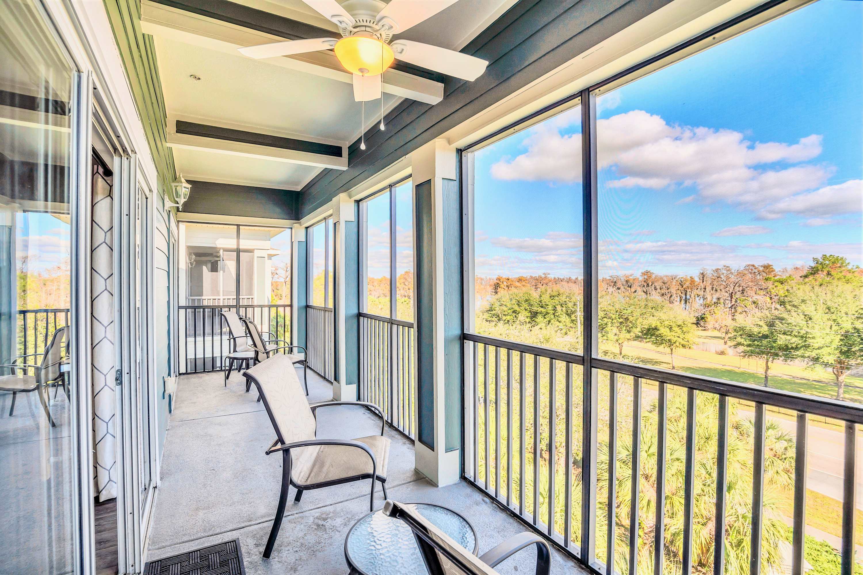 Screened-in balcony with outdoor seating, offering expansive views of lush trees and a distant road under a bright blue sky. A ceiling fan is installed for comfort, and the floor is concrete.
