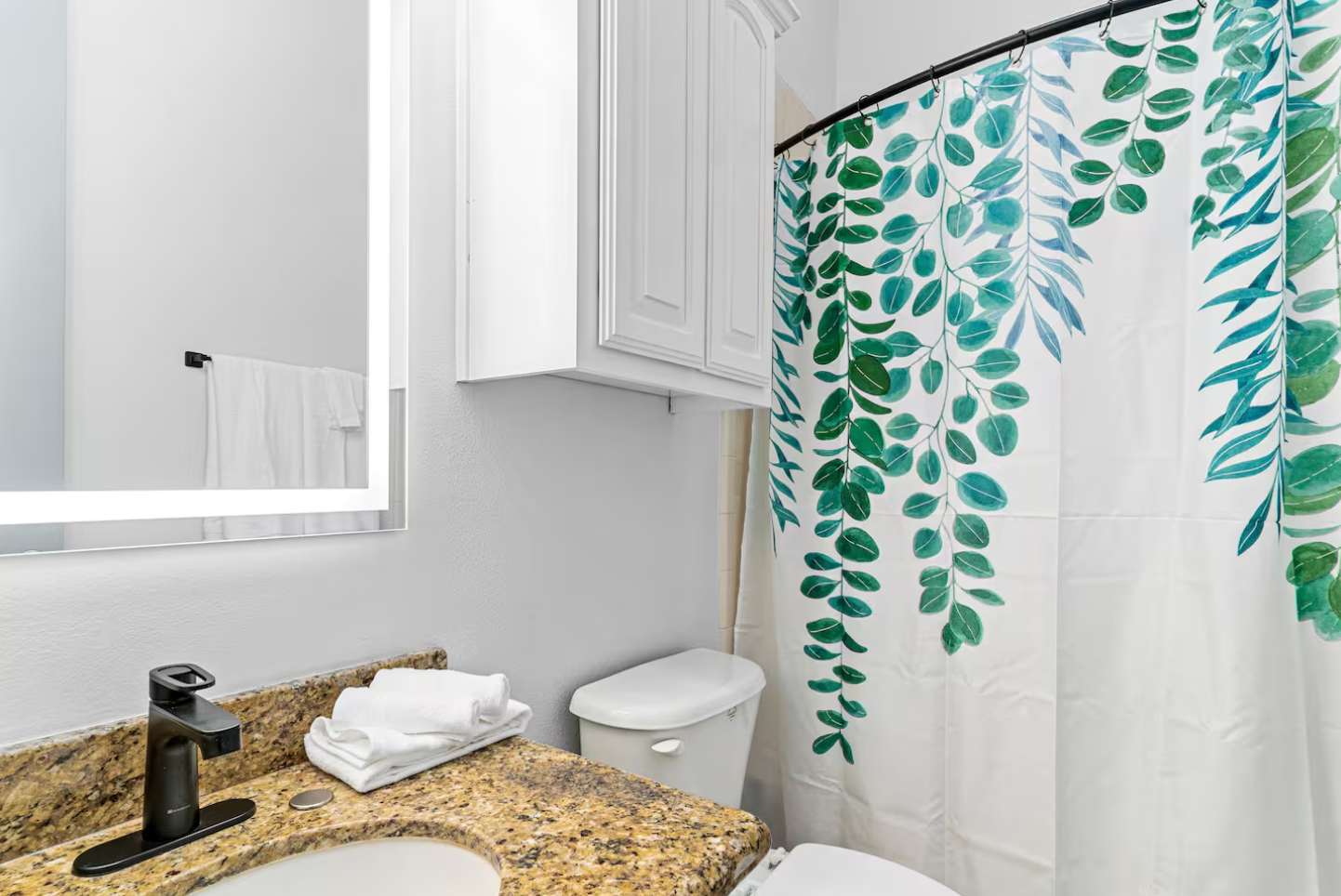 Bathroom featuring a white toilet next to a shower/tub combo with a vibrant green leaf-patterned shower curtain, and a white cabinet mounted above the toilet.