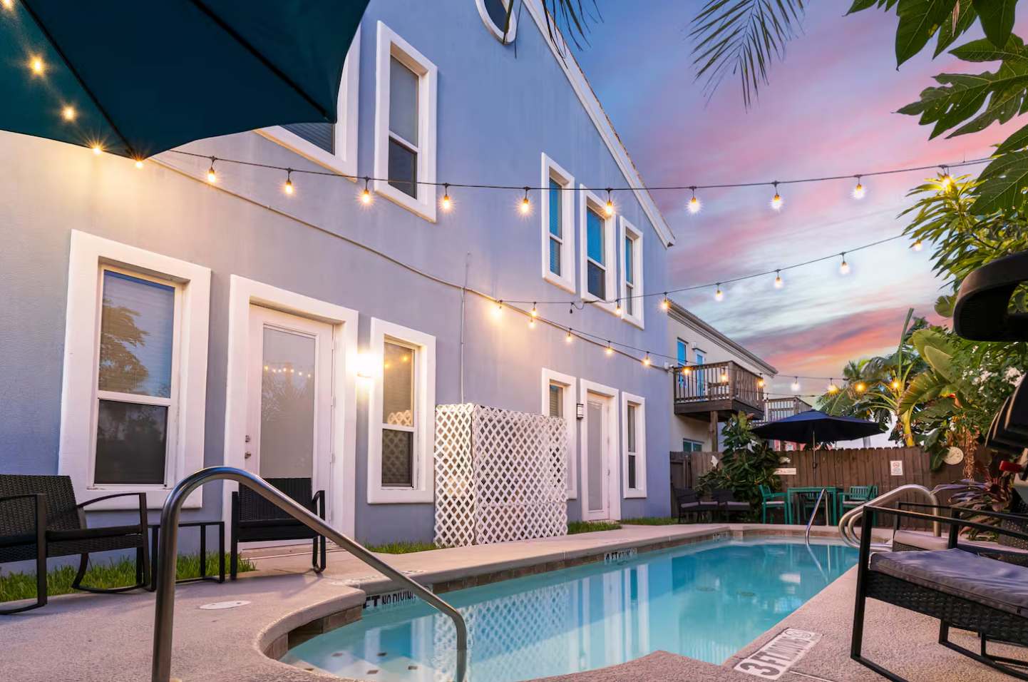 Exterior view of the blue multi-story building from the pool deck, featuring multiple windows and doors, with string lights strung across the patio, creating a warm evening ambiance.