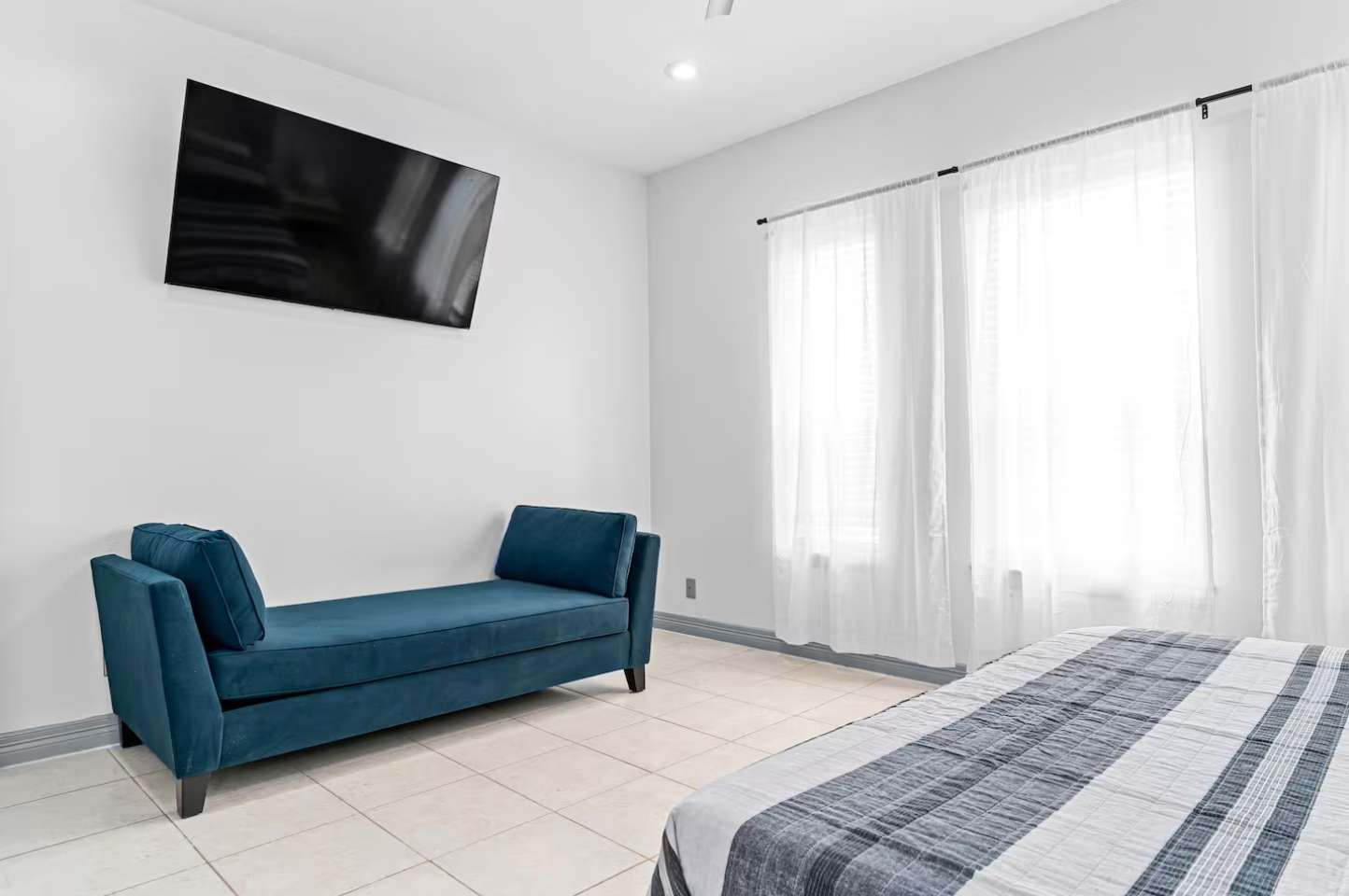 Bright bedroom featuring a large wall-mounted TV above a teal chaise lounge, with light tiled flooring and sheer white curtains covering large windows, and a striped bedspread partially visible.