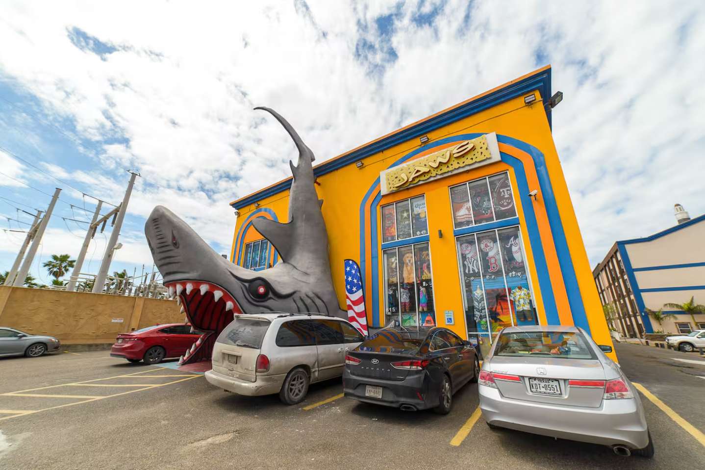 Bright yellow and blue commercial building featuring a large, dramatic shark sculpture bursting from its side. Several cars are parked in the foreground under a partly cloudy sky.
