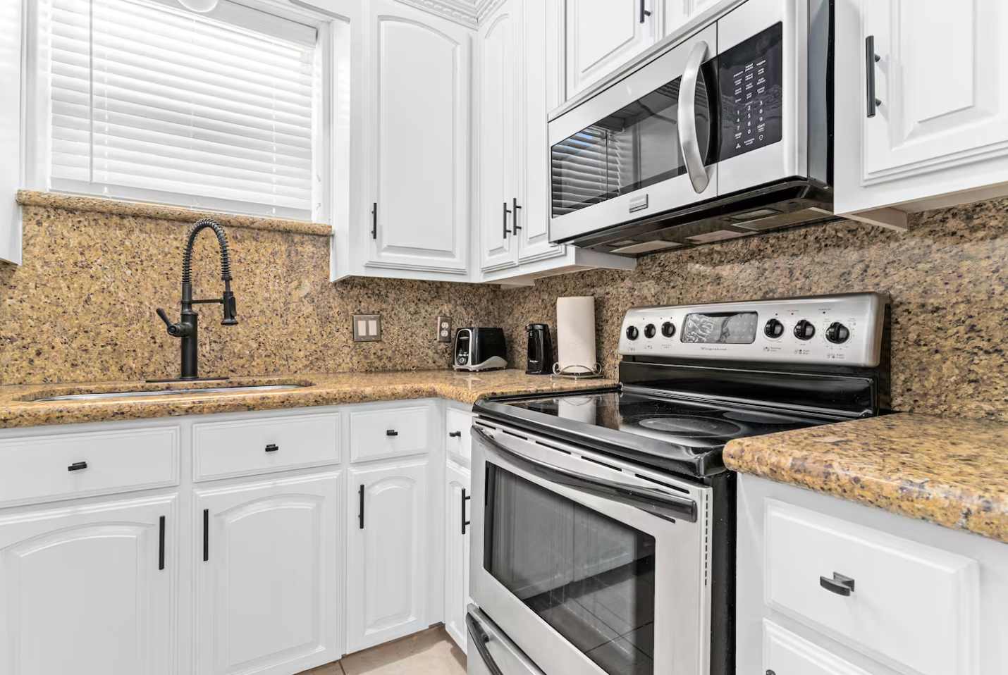 Well-equipped kitchen with a stainless steel electric range and an over-the-range microwave. Dark granite countertops, a black gooseneck faucet, and white cabinets are prominent.