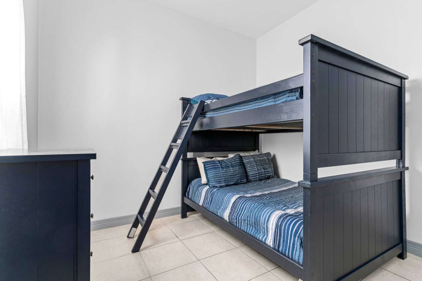 Spacious bedroom showcasing a dark blue wooden bunk bed with a matching dresser, featuring white walls and light tiled flooring, providing a clean and functional sleeping space.