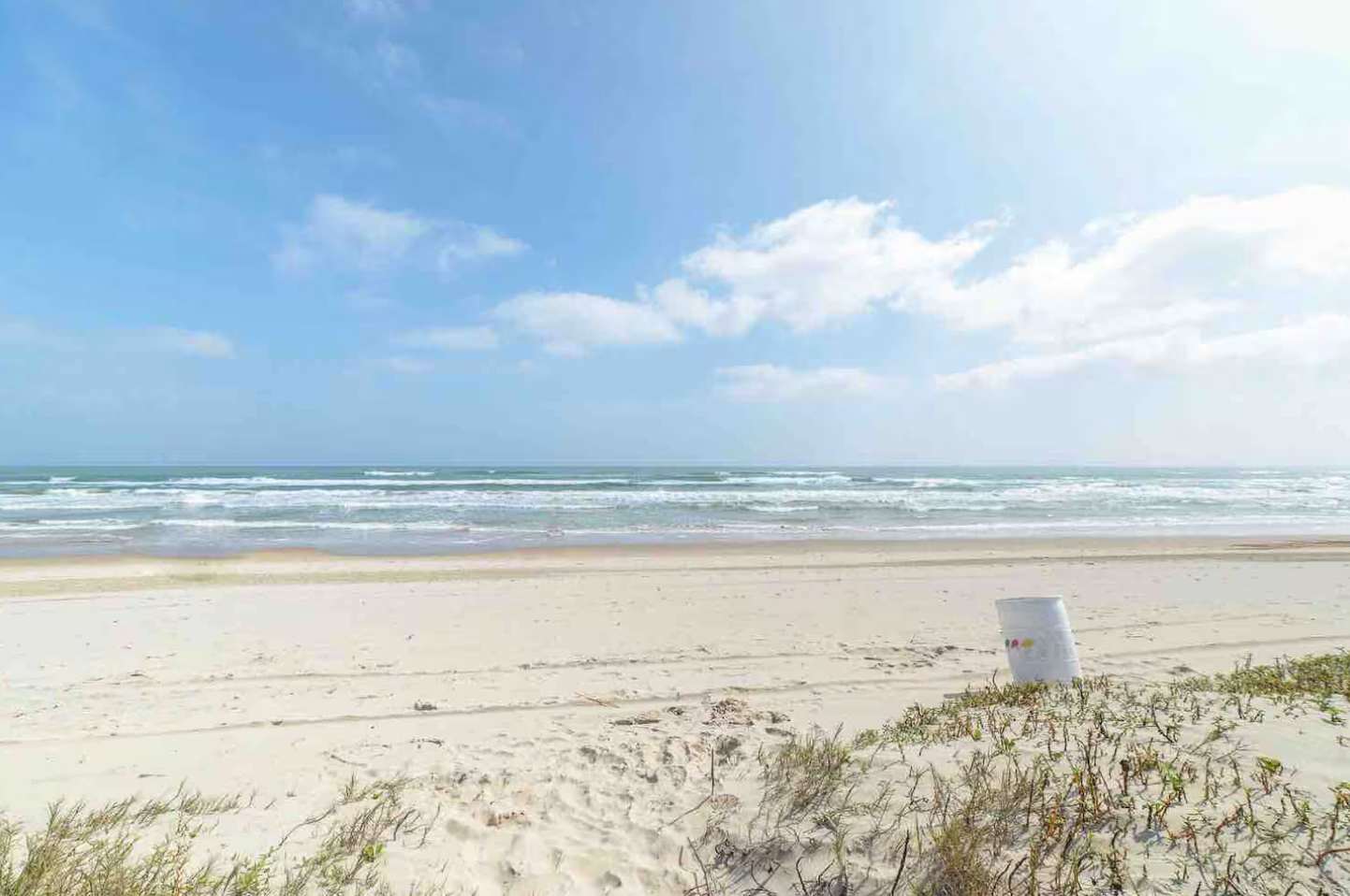 Expansive sandy beach with gentle ocean waves under a bright blue sky with scattered white clouds. Sparse dune vegetation lines the foreground, with a white bucket near the edge.