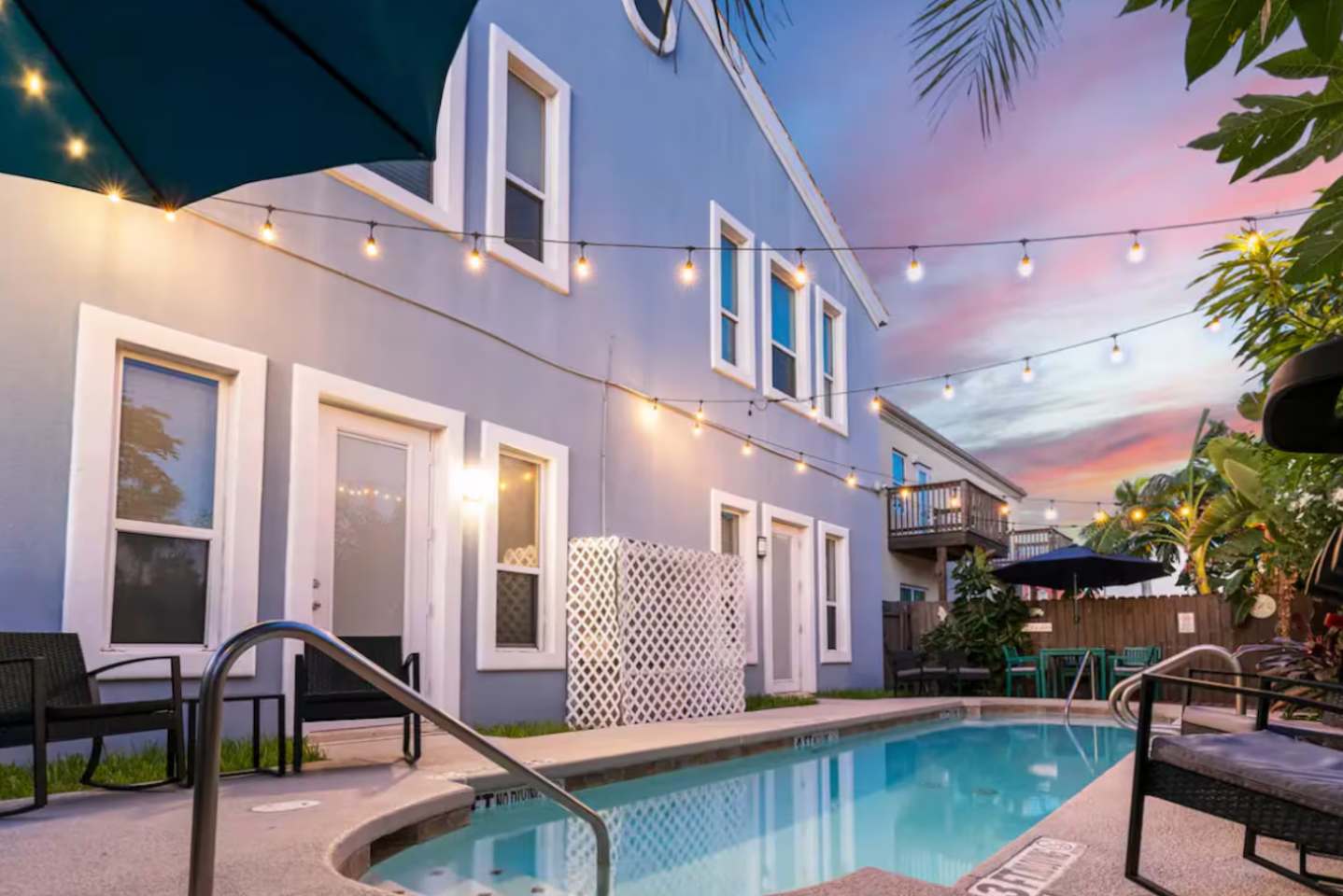 A modern blue building with white trim and multiple windows, illuminated by string lights, overlooks a swimming pool with a railing and outdoor seating. The sky is a vibrant sunset mix of pink and purple.