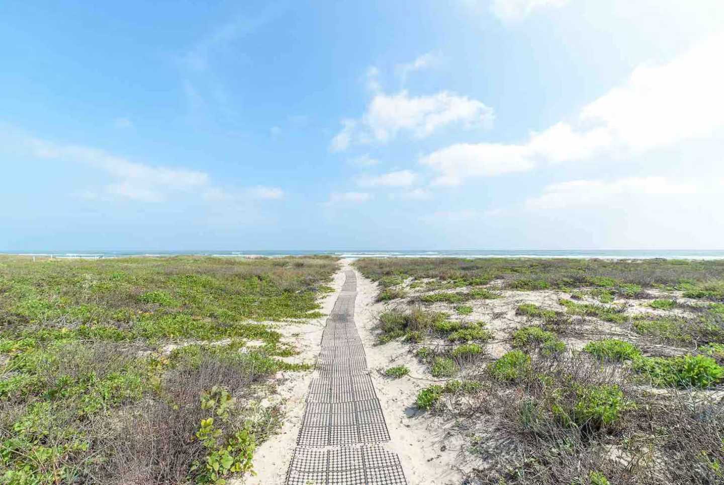 A wooden boardwalk path stretches through sandy dunes covered in green vegetation, leading towards the ocean under a bright blue sky with scattered clouds. The path provides direct access to the beach.