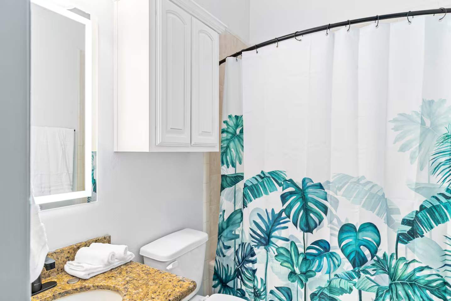 Bathroom featuring a white toilet and a shower/tub combo with a vibrant tropical leaf-patterned shower curtain, complemented by a white cabinet and a modern illuminated mirror.