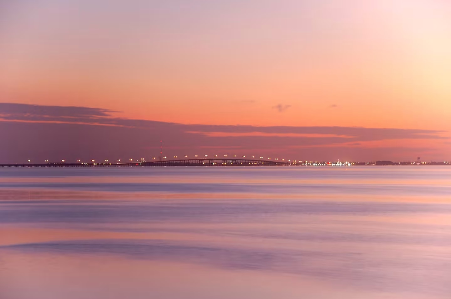 A serene sunset scene over calm water, with a distant bridge illuminated by lights stretching across the horizon. The sky displays soft gradients of pink, orange, and purple.