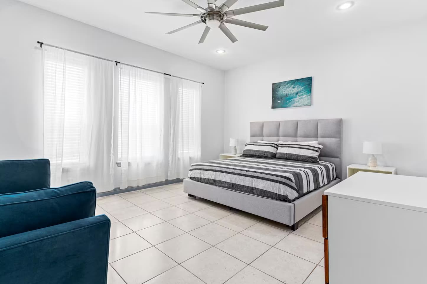 Bright bedroom with a grey upholstered queen bed, white tile floors, and sheer white curtains. A blue armchair and a white dresser are also present, adding functional seating and storage.