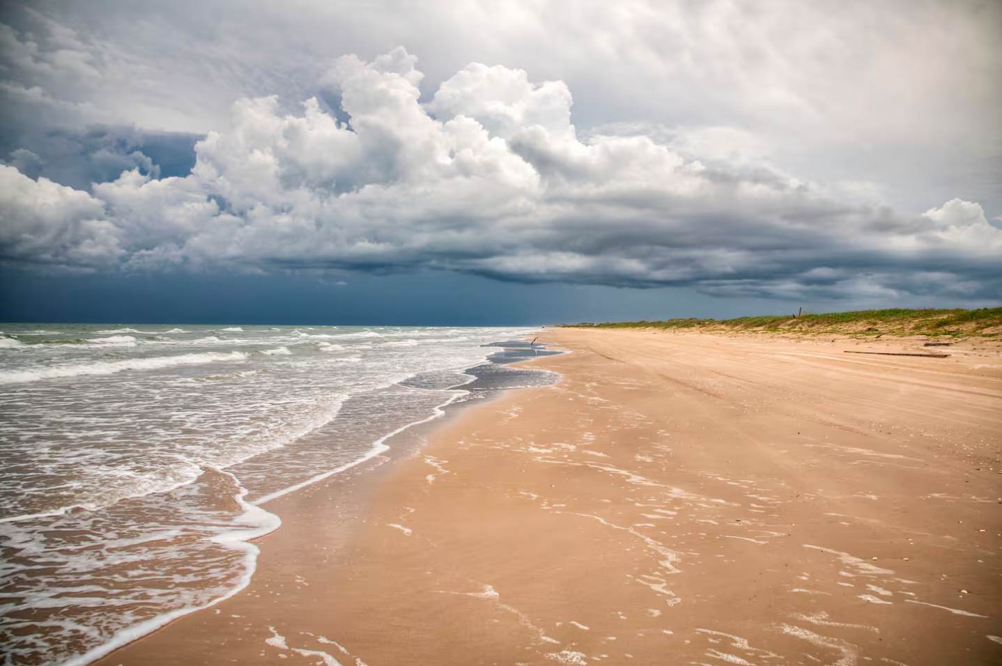 Vast, empty sandy beach with foamy ocean waves receding from the shore under a dramatic, heavily clouded sky. The beach extends into the horizon with distant dunes.