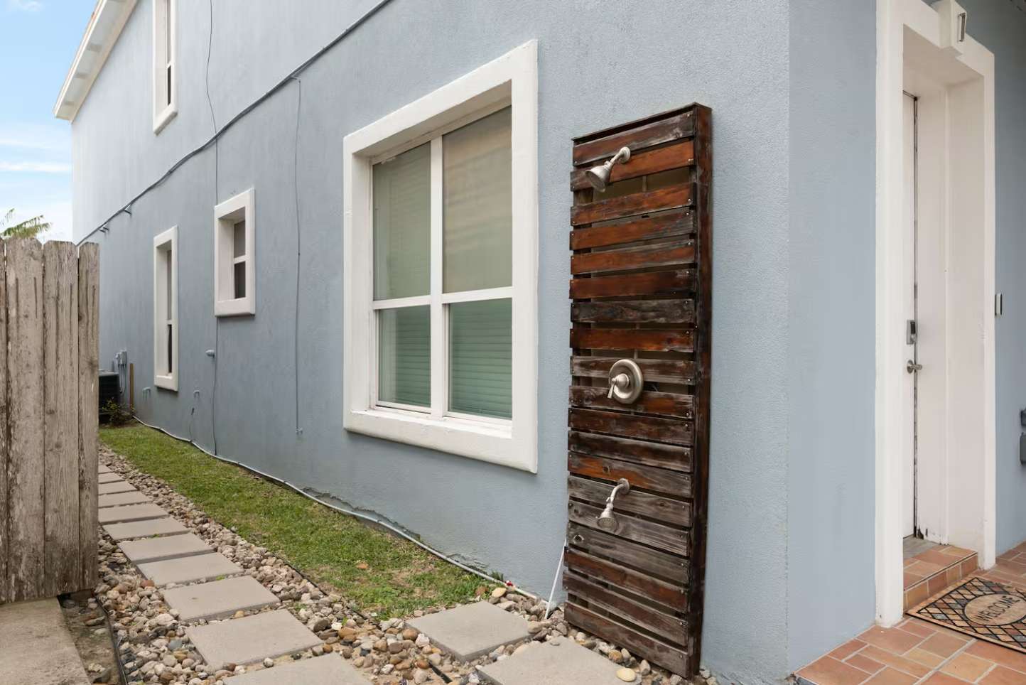 An outdoor shower with a slatted wooden panel is mounted on the light blue exterior wall of the building, next to a white door. A stone path with gravel runs alongside a strip of grass.